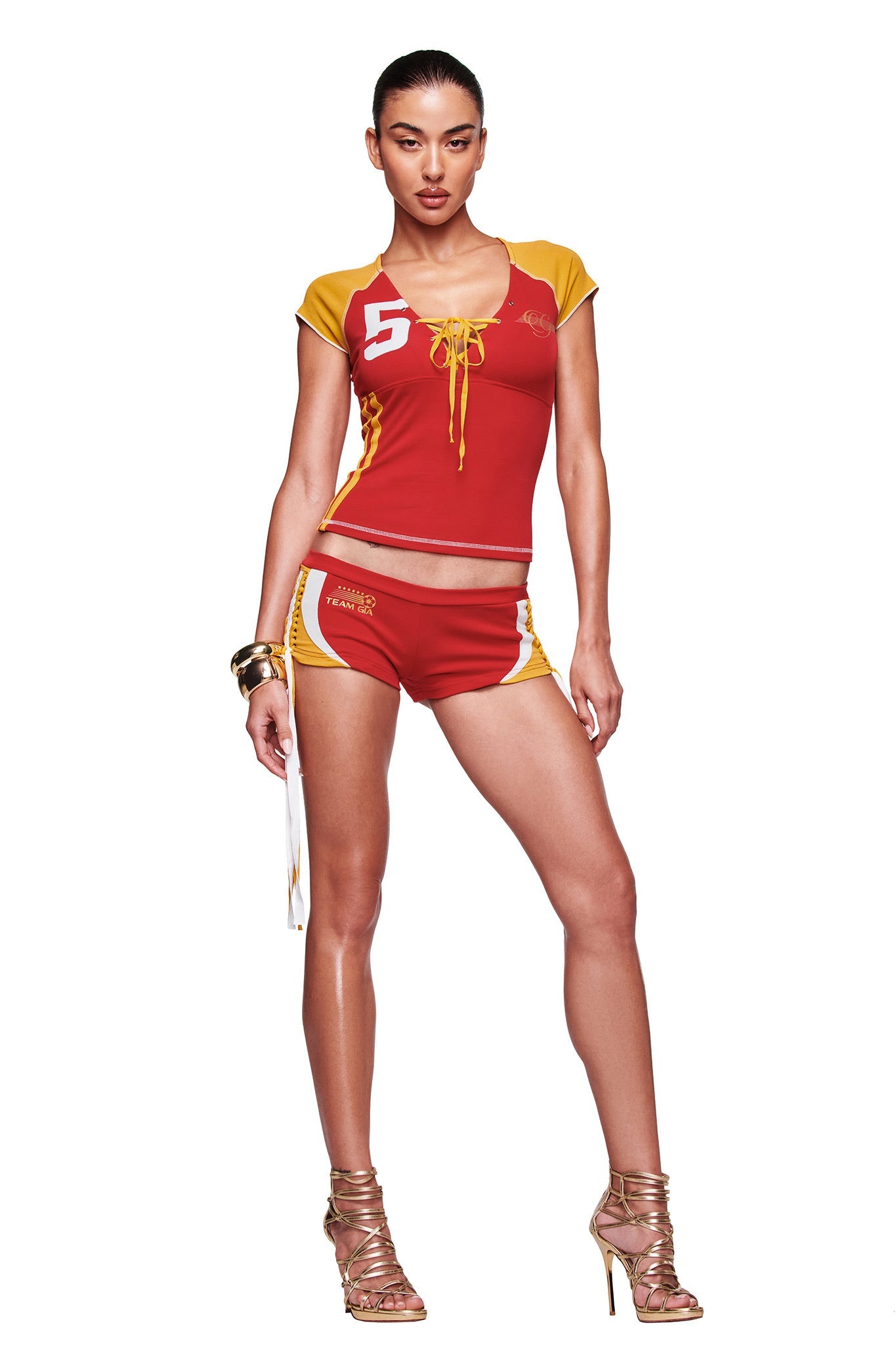 A woman stands confidently in the I.AM.GIA NOVIA MICRO SPORT SHORT - RED, styled with a red and yellow sports top featuring the number 5, gold high-heeled sandals, and gold bracelets, posed against a white background.