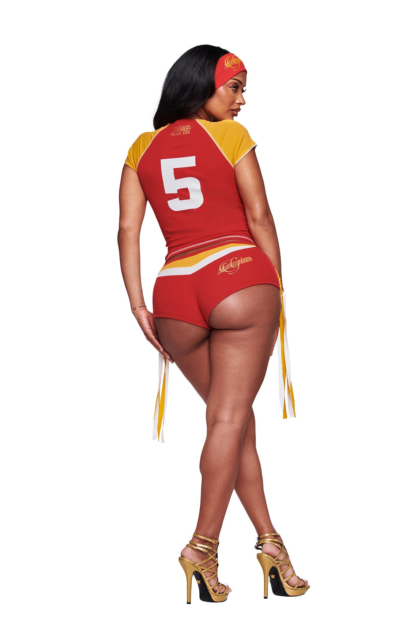 A woman faces away against a white background, wearing I.AM.GIA's NOVIA MICRO SPORT SHORT in red, a sporty top with the number 5, gold high heels, and a matching headband.