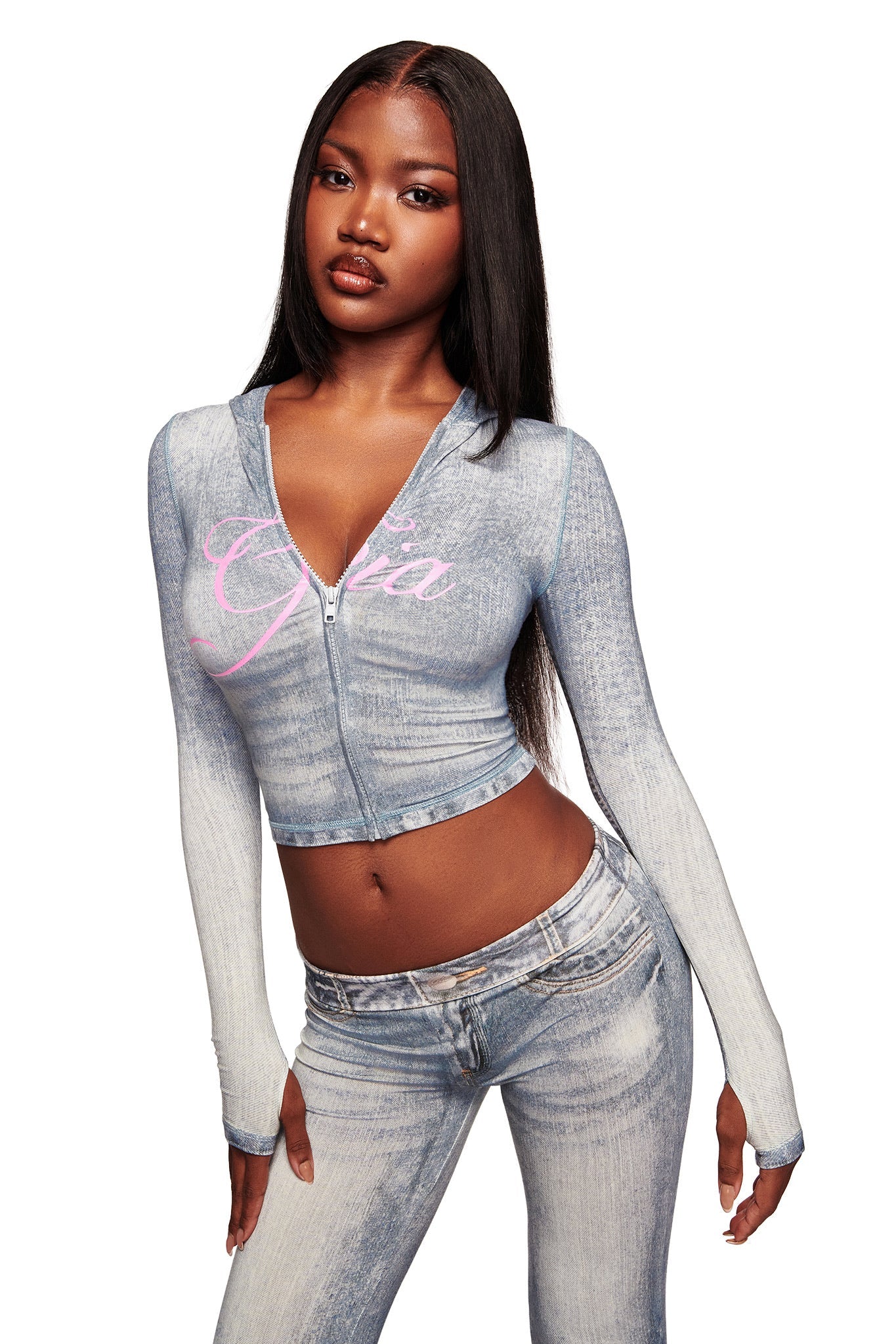 A woman with long straight hair wears the I.AM.GIA BLARE HOODIE - DENIM TROMPE L'OEIL, a light blue cropped hoodie featuring "GIA" in pink script, paired with matching high-waisted jeans, posing confidently against a white background.