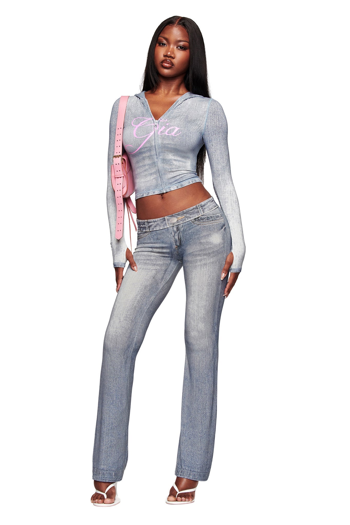 A woman stands confidently in the I.AM.GIA BLARE HOODIE - DENIM TROMPE L'OEIL and matching flared jeans, paired with clear heels and a pink shoulder bag. Her long, straight hair complements the hoodie’s pink "GIA" script detail.