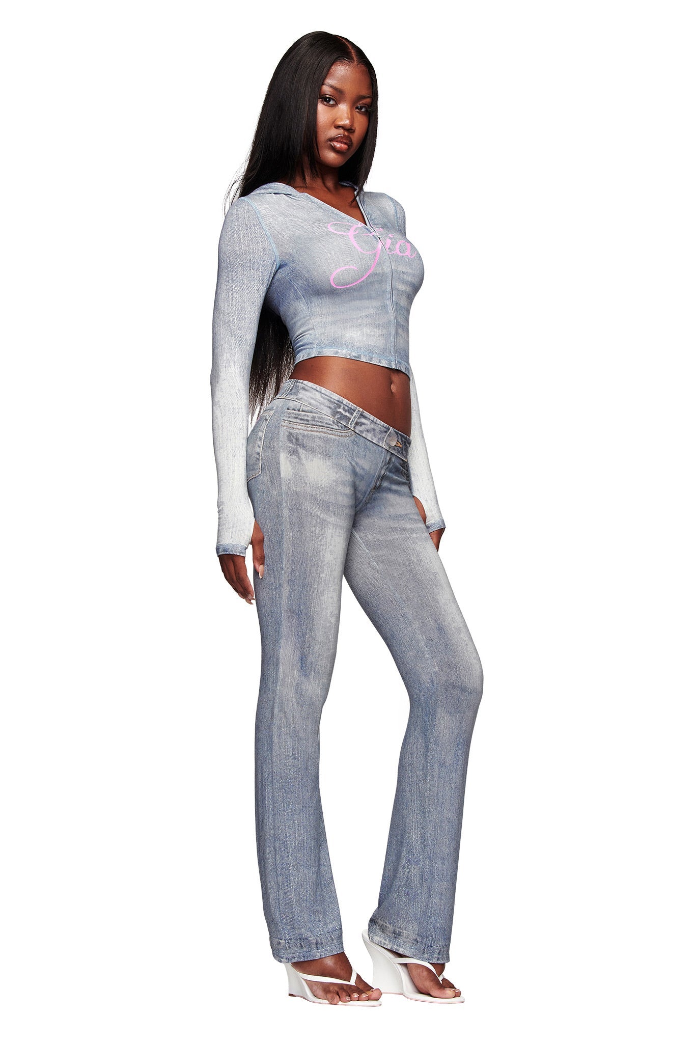 A woman stands against a white background wearing I.AM.GIA's BLARE TRACKPANT - DENIM TROMPE L'OEIL and a matching long-sleeve crop top with “Grace” in pink, styled with white open-toe heels and long straight hair.