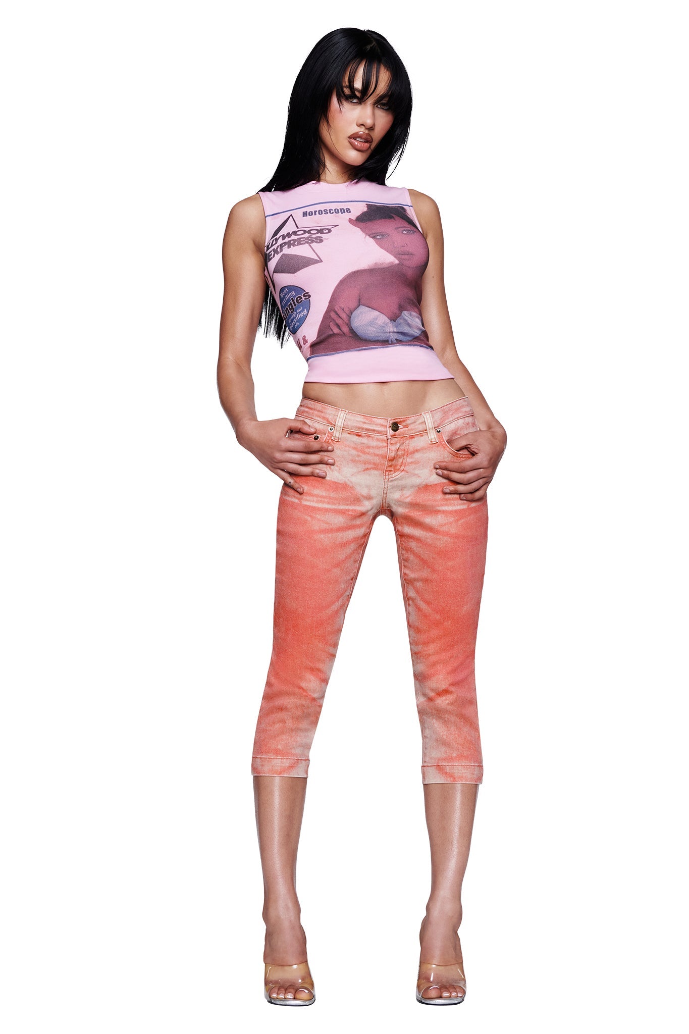 A woman poses against a white background wearing the I.AM.GIA SUNSET CAPRI JEAN in Peach, featuring an ombré wash and crafted from stretch denim, styled with a sleeveless pink graphic crop top and clear high-heeled shoes.