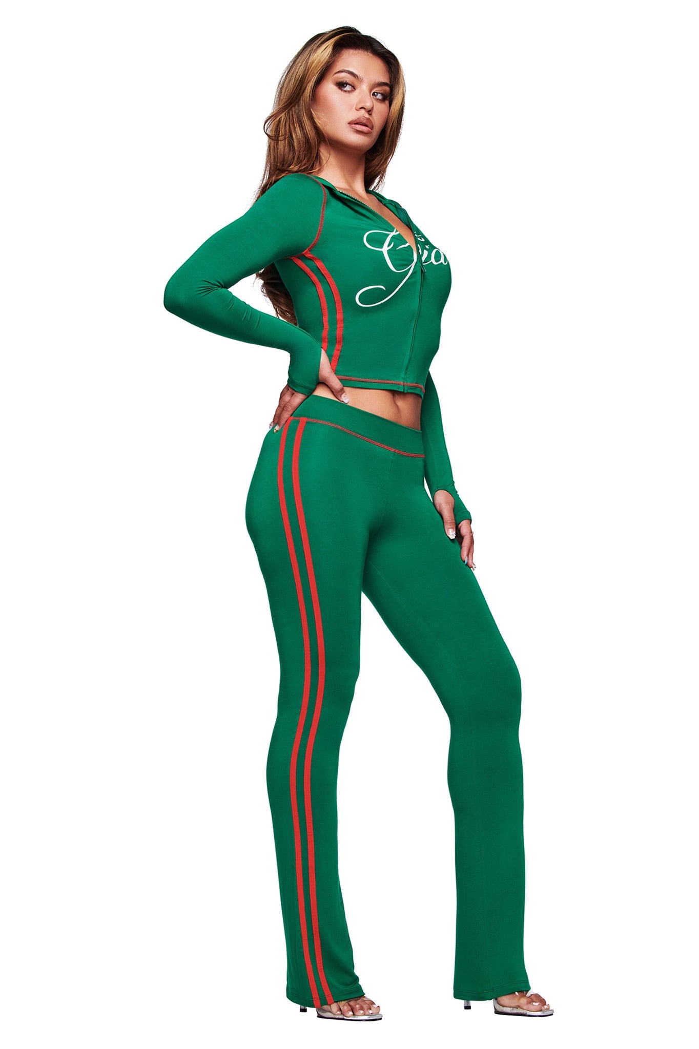 A woman wears the I.AM.GIA BLARE SPORT TRACKPANT in green, featuring red side stripes and a flared fit. She pairs them with a long-sleeve graphic top and high heels for a sporty-chic look.