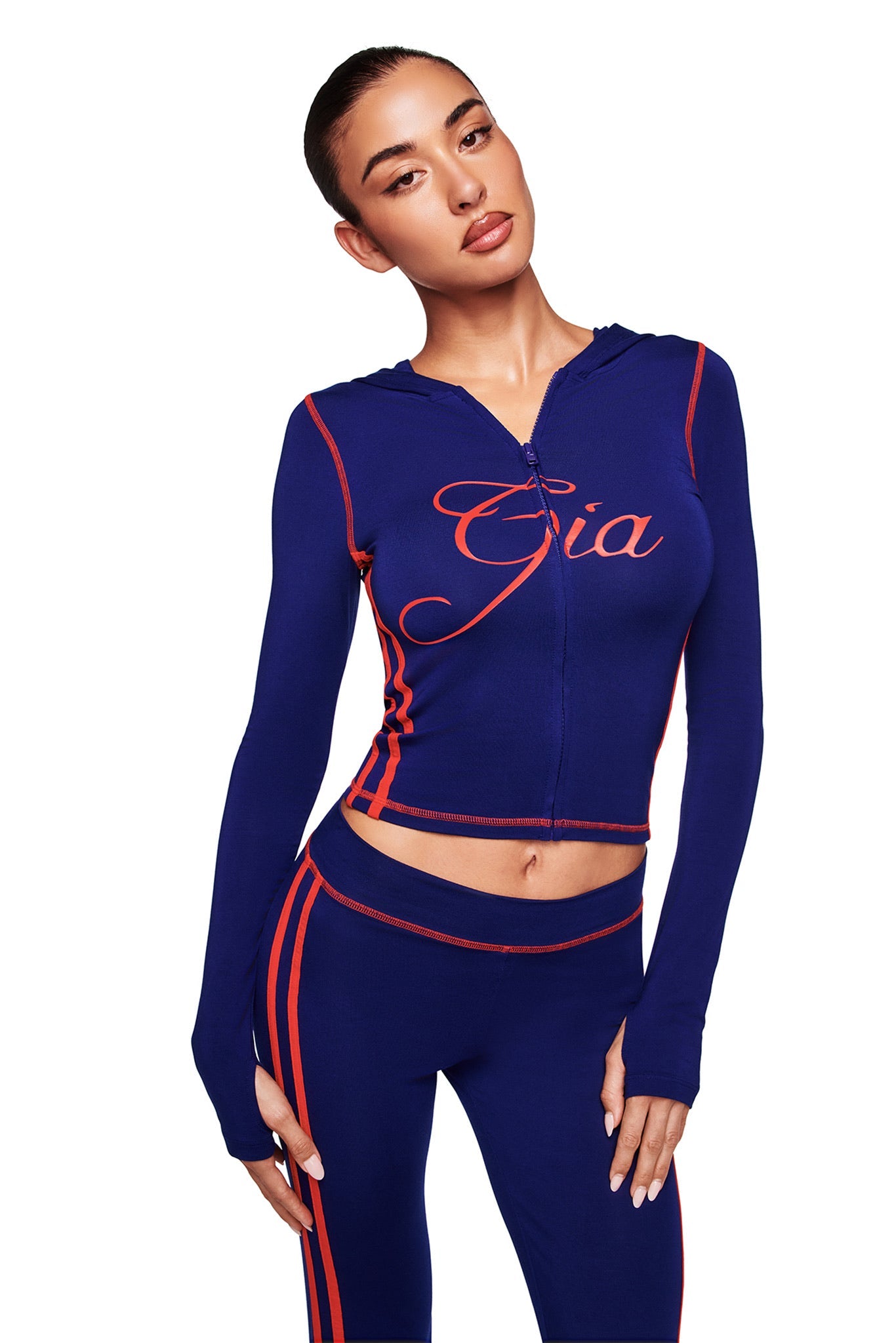 A woman models the I.AM.GIA BLARE TWO-PIECE SPORT HOODIE in navy, featuring red trim and "Gia" on the front, with thumb-hole sleeves and matching blue pants with red side stripes, posing against a white background.