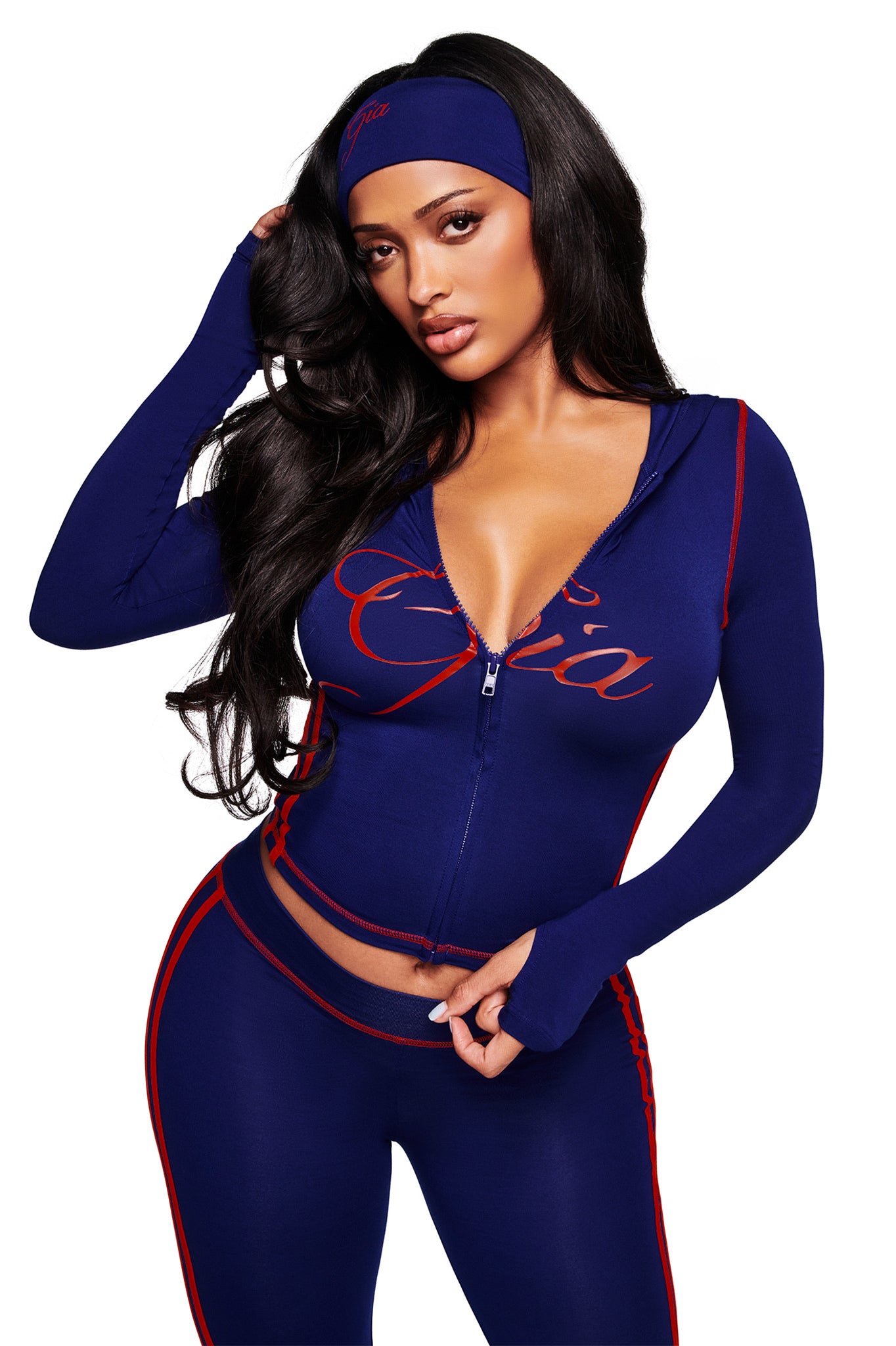 A woman with long dark hair wears the I.AM.GIA BLARE SPORT HOODIE AND HEADBAND in navy, featuring red accents, script text, a front zipper, and thumb-hole detail as she poses with her hand touching her head.