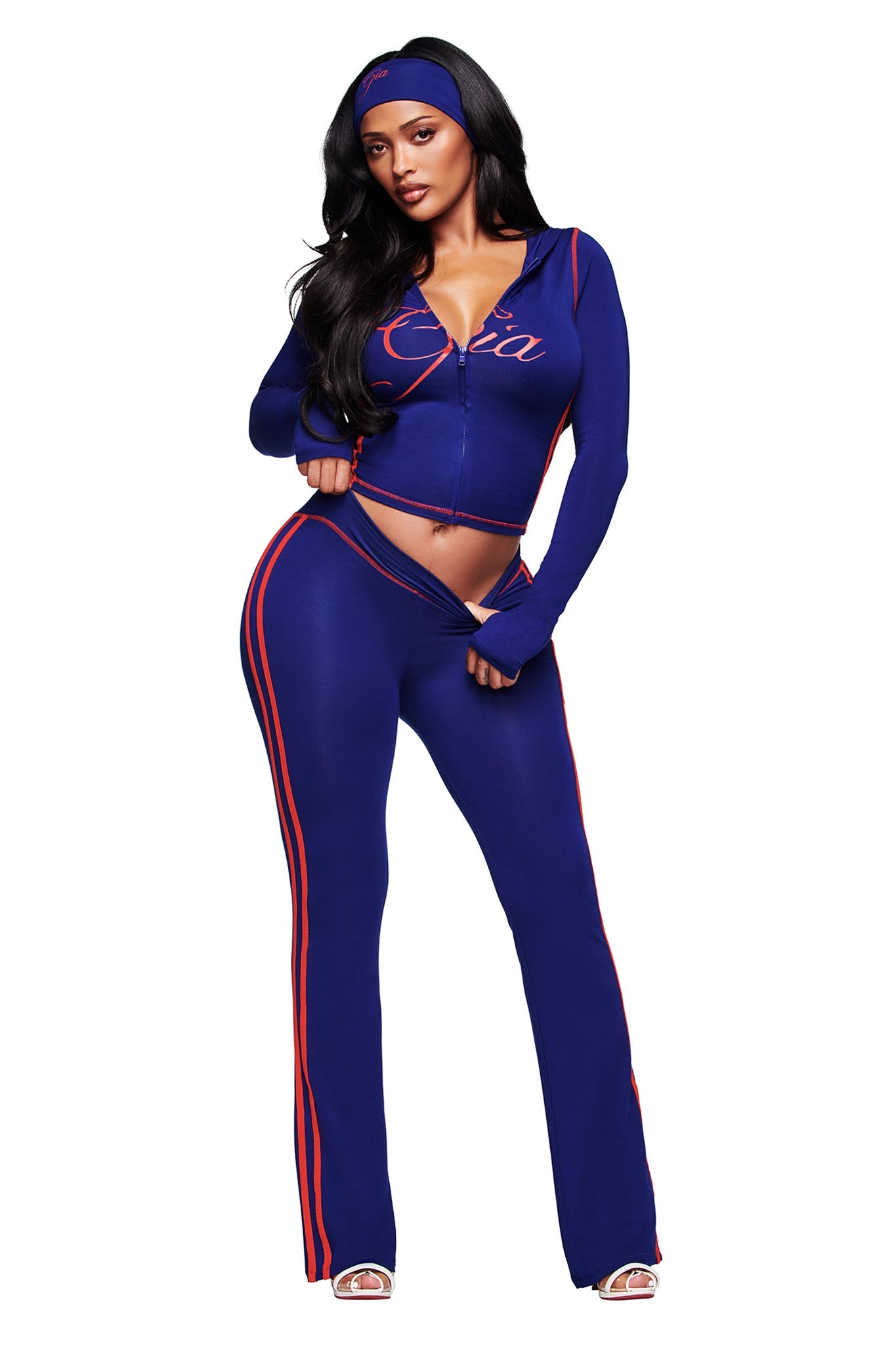 A woman poses in the I.AM.GIA BLARE TWO-PIECE SPORT HOODIE - NAVY, featuring matching pants, thumb-hole cut-outs, and a modern fit. She accessorizes with a headband and open-toe heels against a white background.