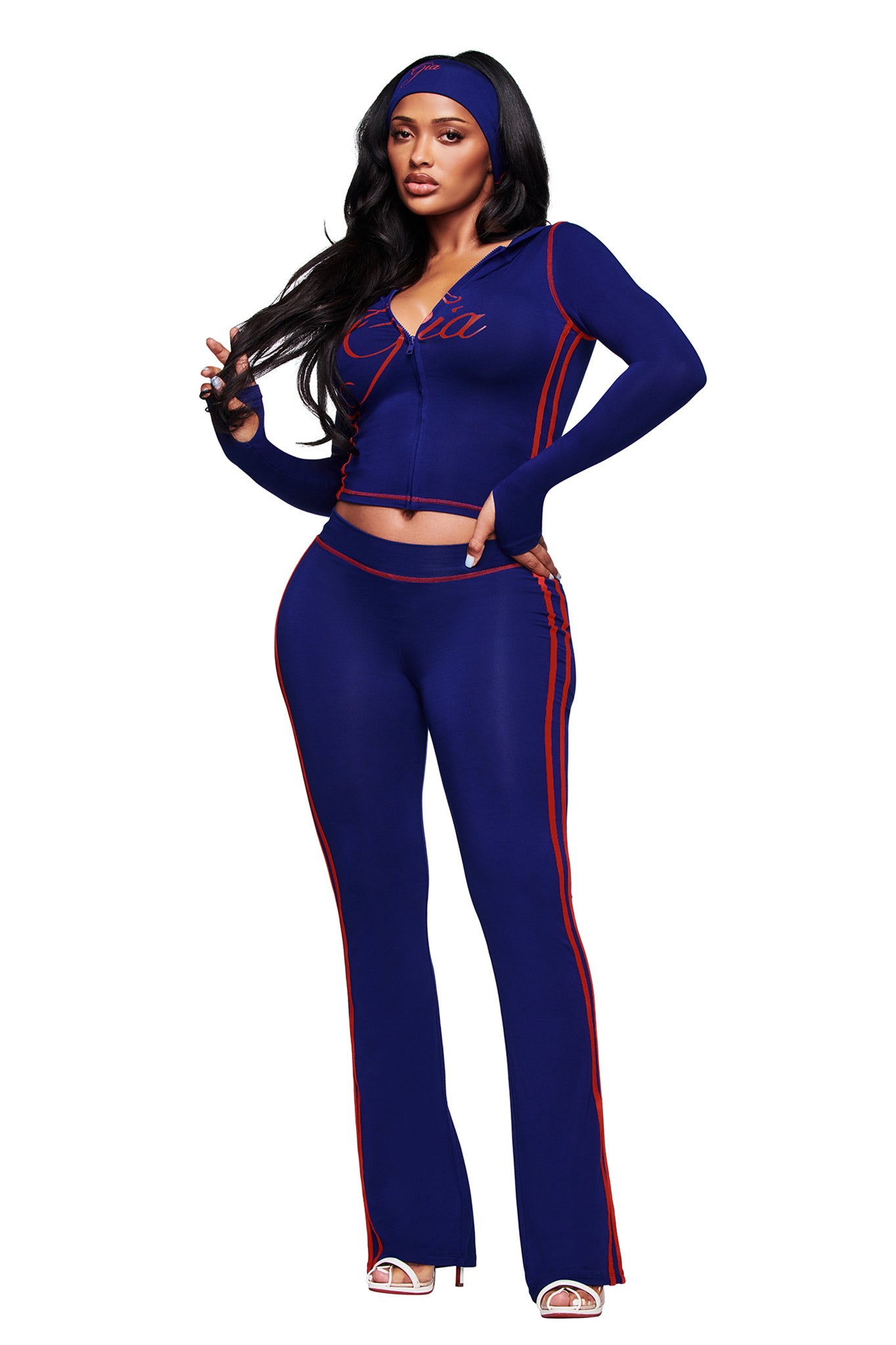 A woman poses against a white background wearing I.AM.GIA’s BLARE SPORT TRACKPANT in navy—a fitted, buttery-soft sporty style—paired with a matching jacket with red stripes and "Coca," plus a headband and heeled sandals.