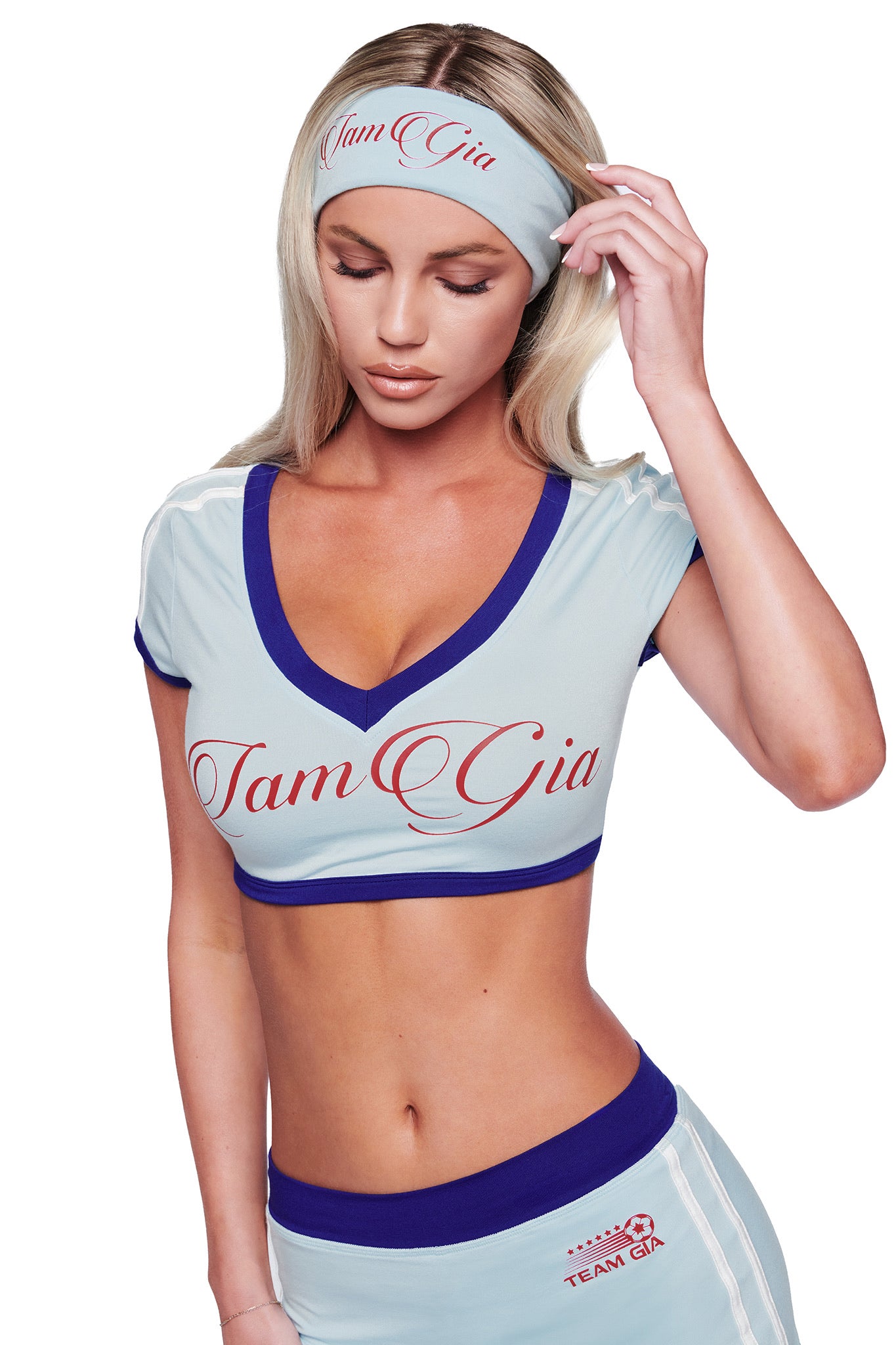 A woman with long blonde hair wears a light blue crop top and the I.AM.GIA BLARE SPORT HEADBAND in Baby Blue, both featuring "Team Gia" in red script. She looks down, touching the stylish headband with one hand.