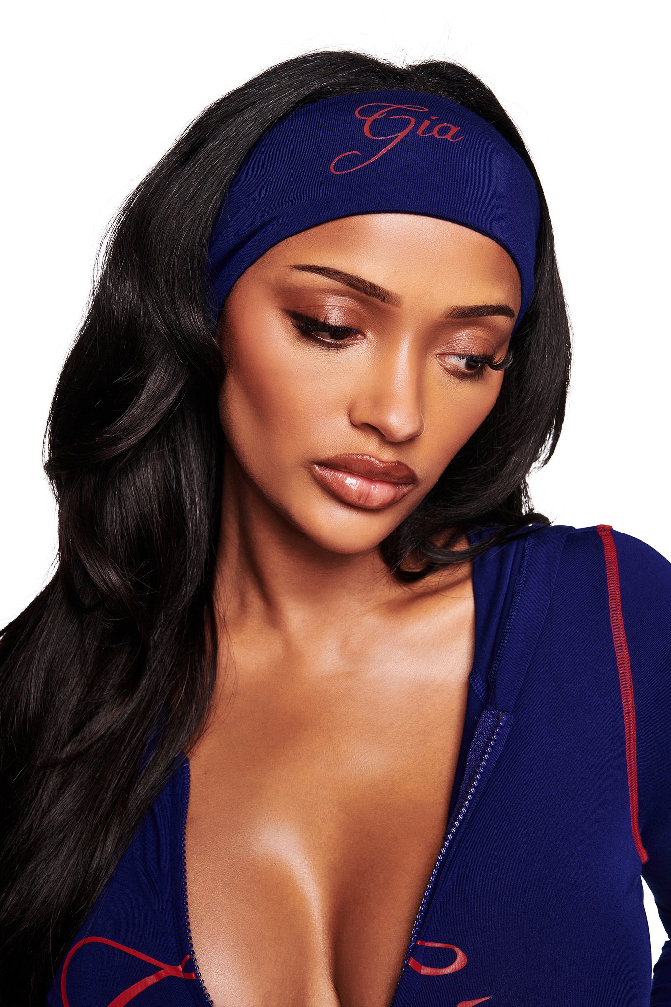 A woman with long, wavy dark hair wears the I.AM.GIA BLARE SPORT HEADBAND in navy, embroidered with "Gia" in red. She pairs it with a matching blue top with red accents, gazing downward with a soft, thoughtful expression.