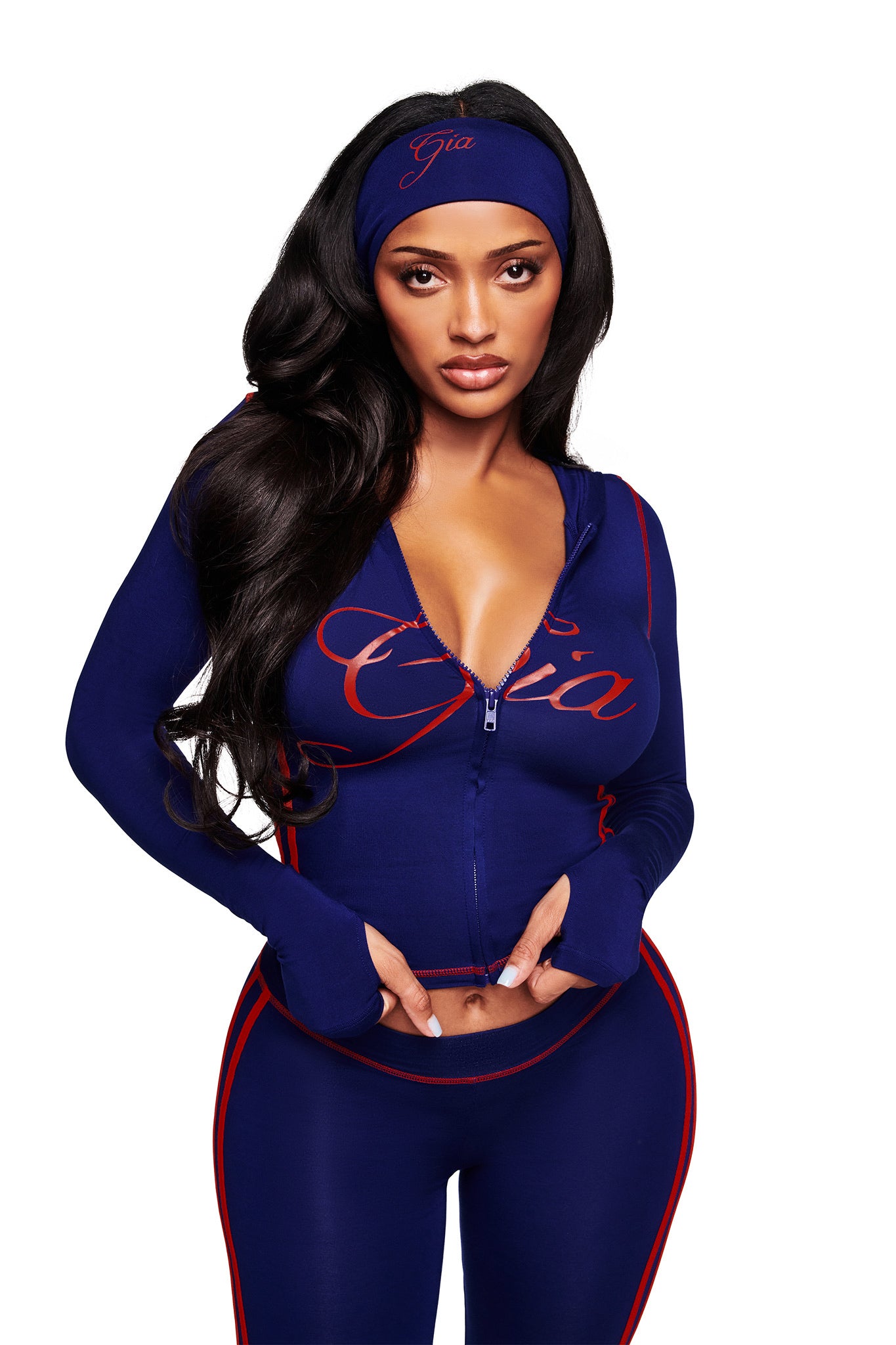 A woman with long dark hair wears a matching navy blue athletic outfit with red trim and the BLARE SPORT HEADBAND - NAVY by I.AM.GIA. She looks at the camera, slightly pulling up her zip-up top against a white background.