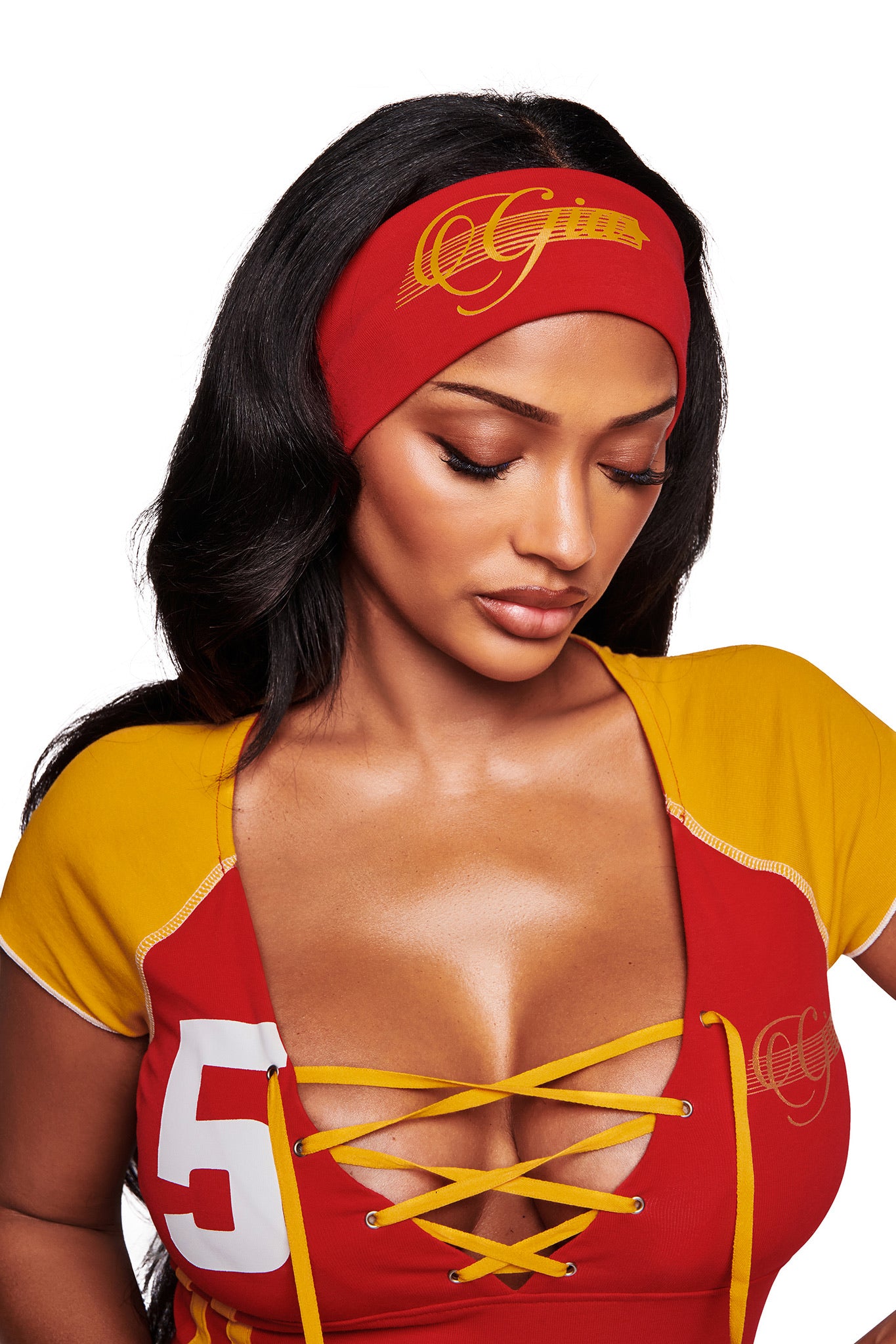 A woman with long dark hair wears the I.AM.GIA NOVIA SPORT HEADBAND - RED and a red and yellow lace-up top with "5" and "OG" on it. She looks down with a neutral expression.