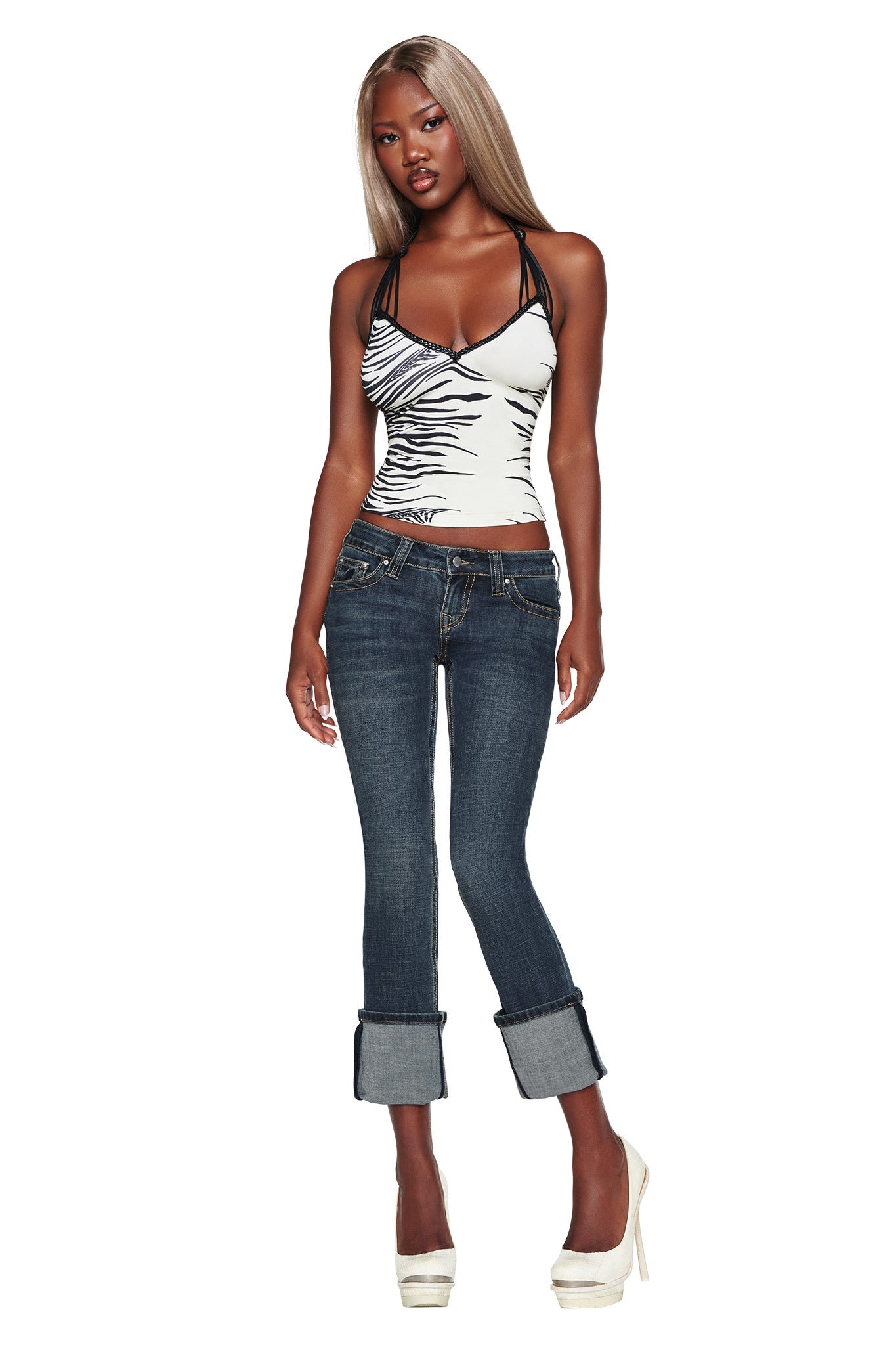 A woman with long blonde hair wears a zebra print tank top, the I.AM.GIA BIANKA JEAN in blue—featuring a slim straight-leg and low-rise fit—and white high heels, standing against a white background.