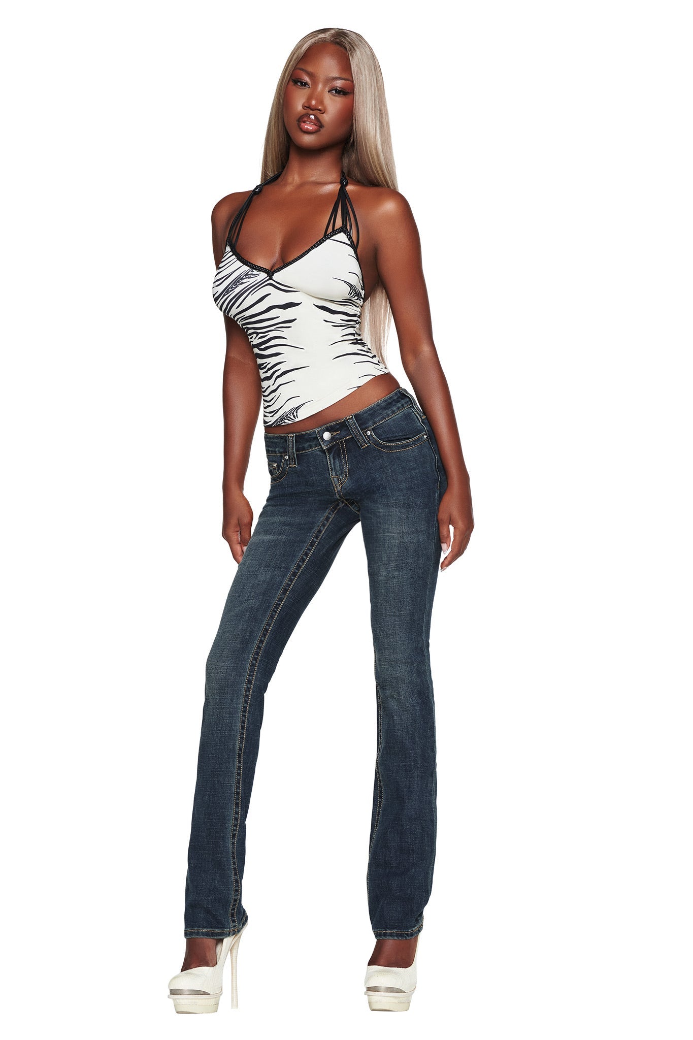 A woman with long blonde hair wears a white and black camisole, the I.AM.GIA BIANKA JEAN in blue featuring a slim straight-leg and low-rise fit, and white platform heels. She stands confidently against a white background.
