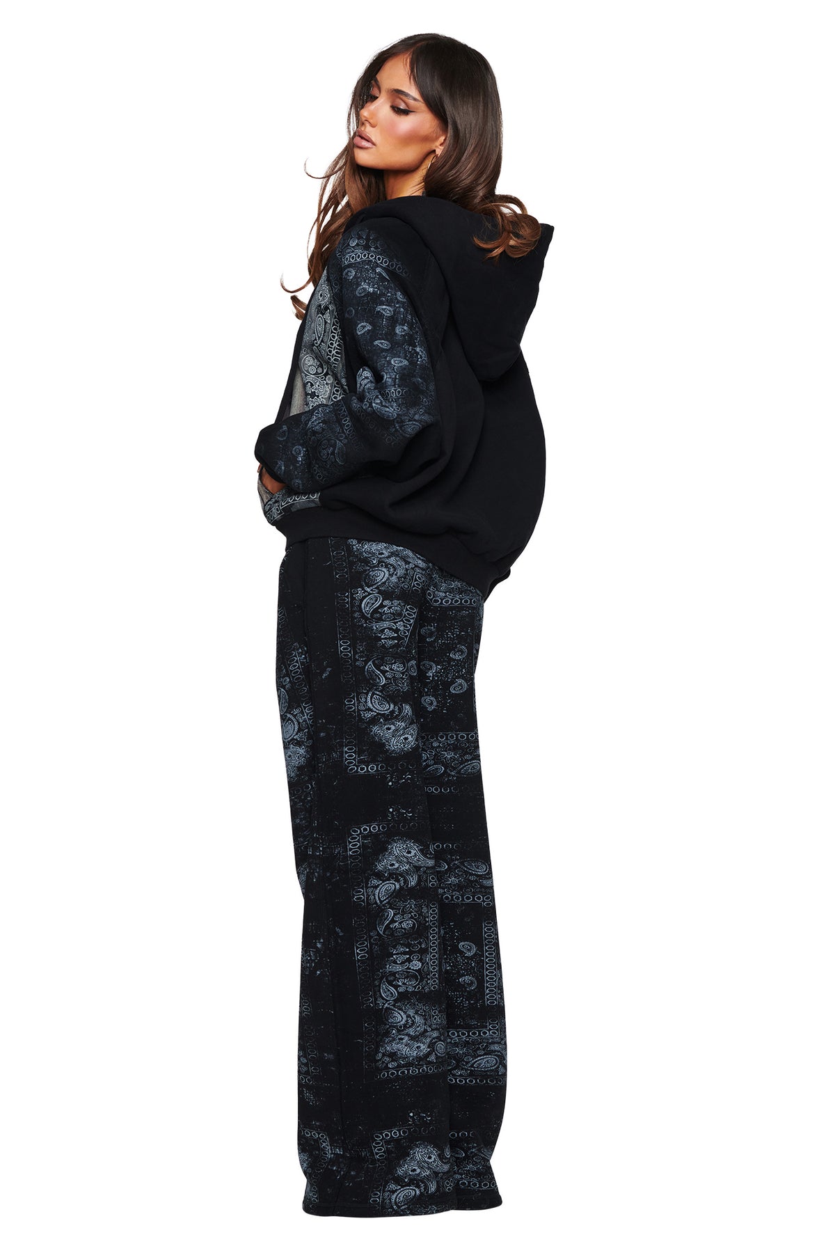 A woman stands in a side pose wearing the I.AM.GIA T-REX ZIP THRU HOODIE - BLACK BANDANA, featuring patterned sleeves, paired with matching wide-leg pants in a blue paisley design for an effortless street style look. She has long, wavy hair and gazes over her shoulder against a white background.