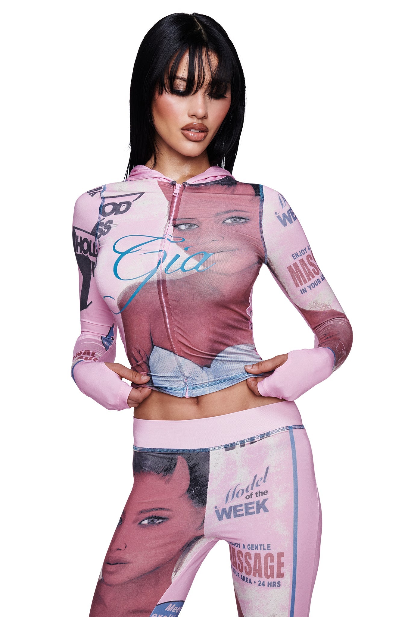 A woman with straight, dark hair and bangs poses with her hands on her hips against a white background, wearing the I.AM.GIA T-REX BLARE HOODIE in pink—a zip-up cropped style—paired with matching leggings featuring vintage magazine-inspired prints and a bold face graphic.
