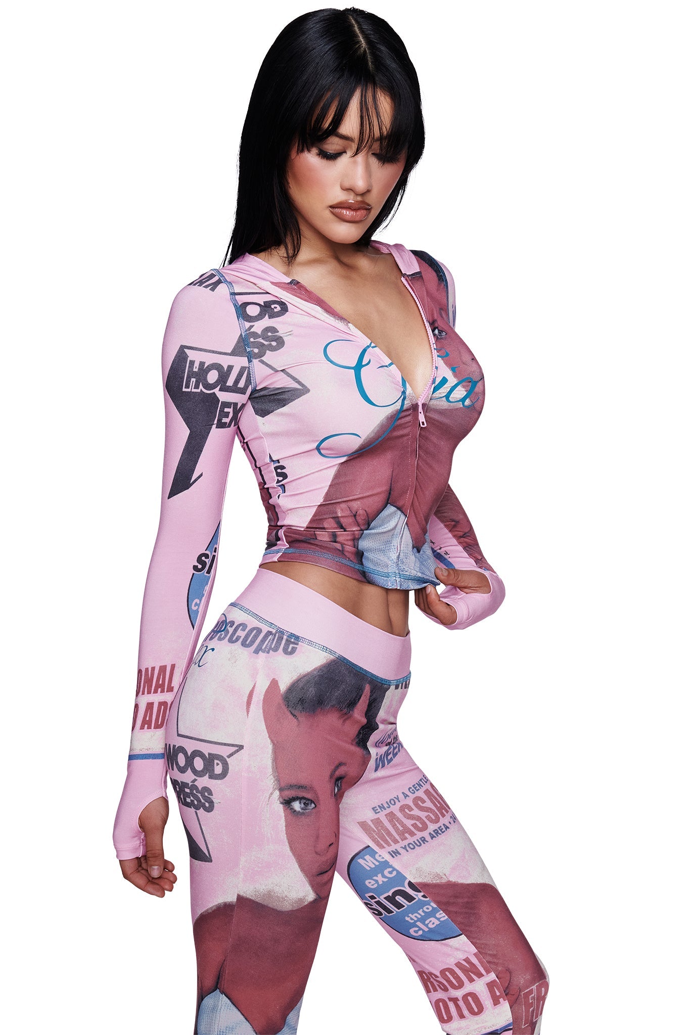 A woman with black hair wears the I.AM.GIA T-REX BLARE TRACKPANT in pink, a fitted, long-sleeve outfit adorned with vintage comic-style illustrations and bold T-REX pink print. She poses, looking down against a plain white background.