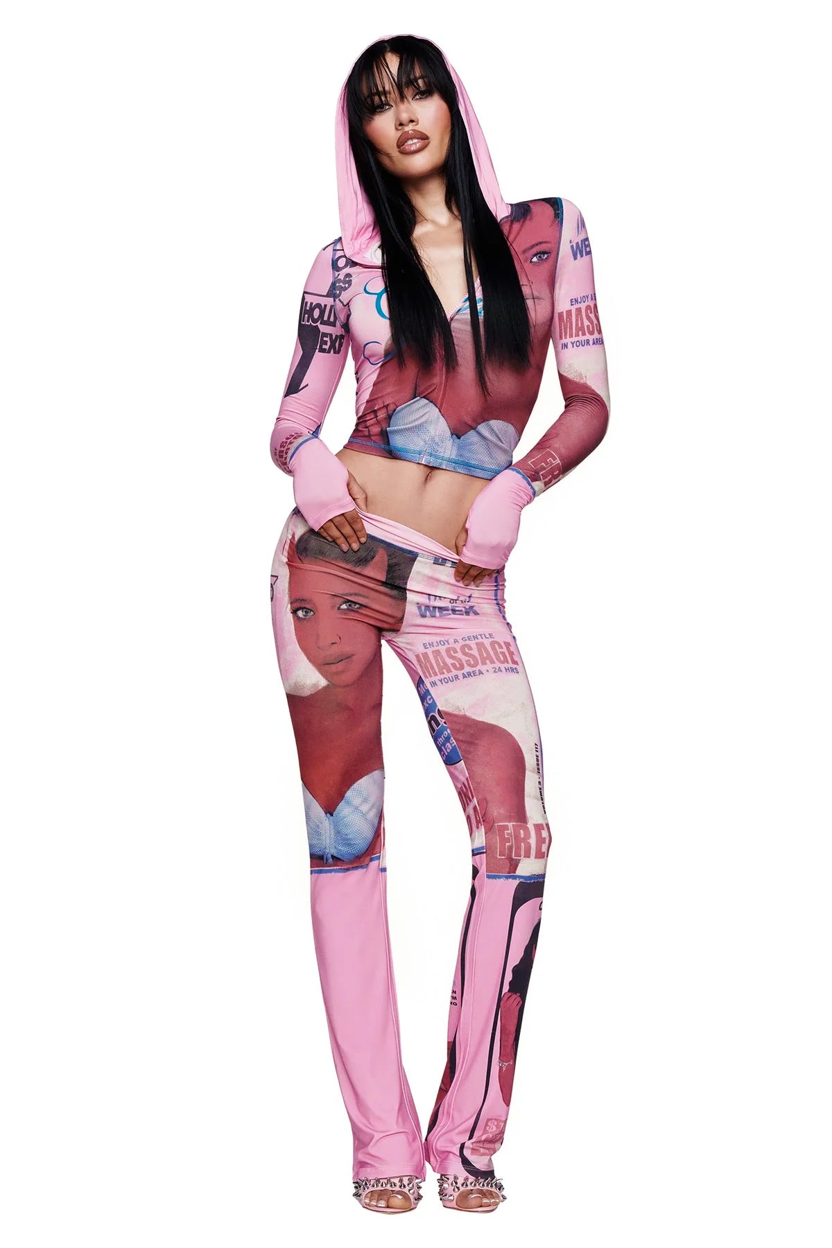 A woman with long black hair and fringe is wearing the I.AM.GIA T-REX BLARE HOODIE in pink, a hooded outfit featuring cartoon-style faces and magazine-inspired text. She stands with one leg bent and her hands in gloves.