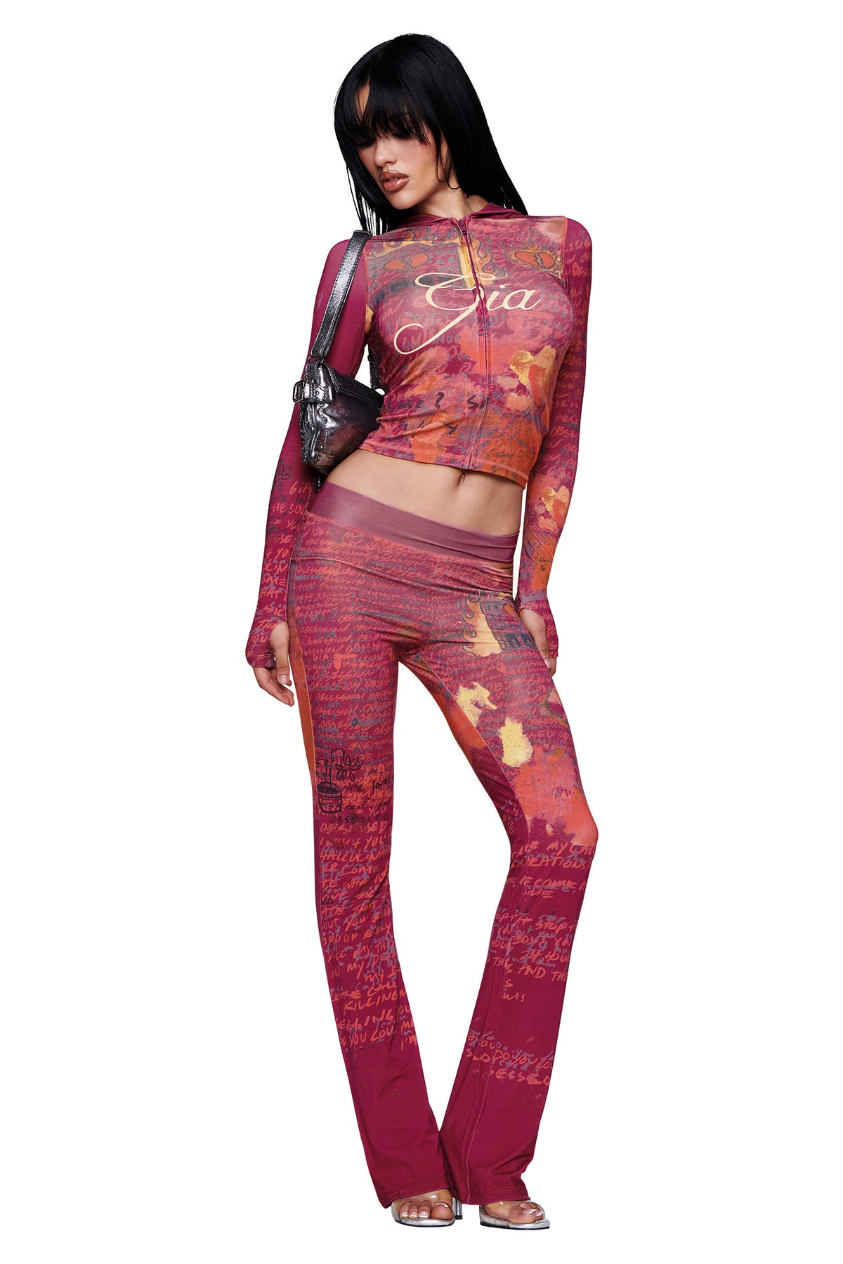 A woman poses against a white background wearing the I.AM.GIA T-REX BLARE HOODIE in red—a fitted, long-sleeve hoodie featuring abstract patterns and logo text—paired with matching pants, styled with a small shoulder bag and heeled sandals.