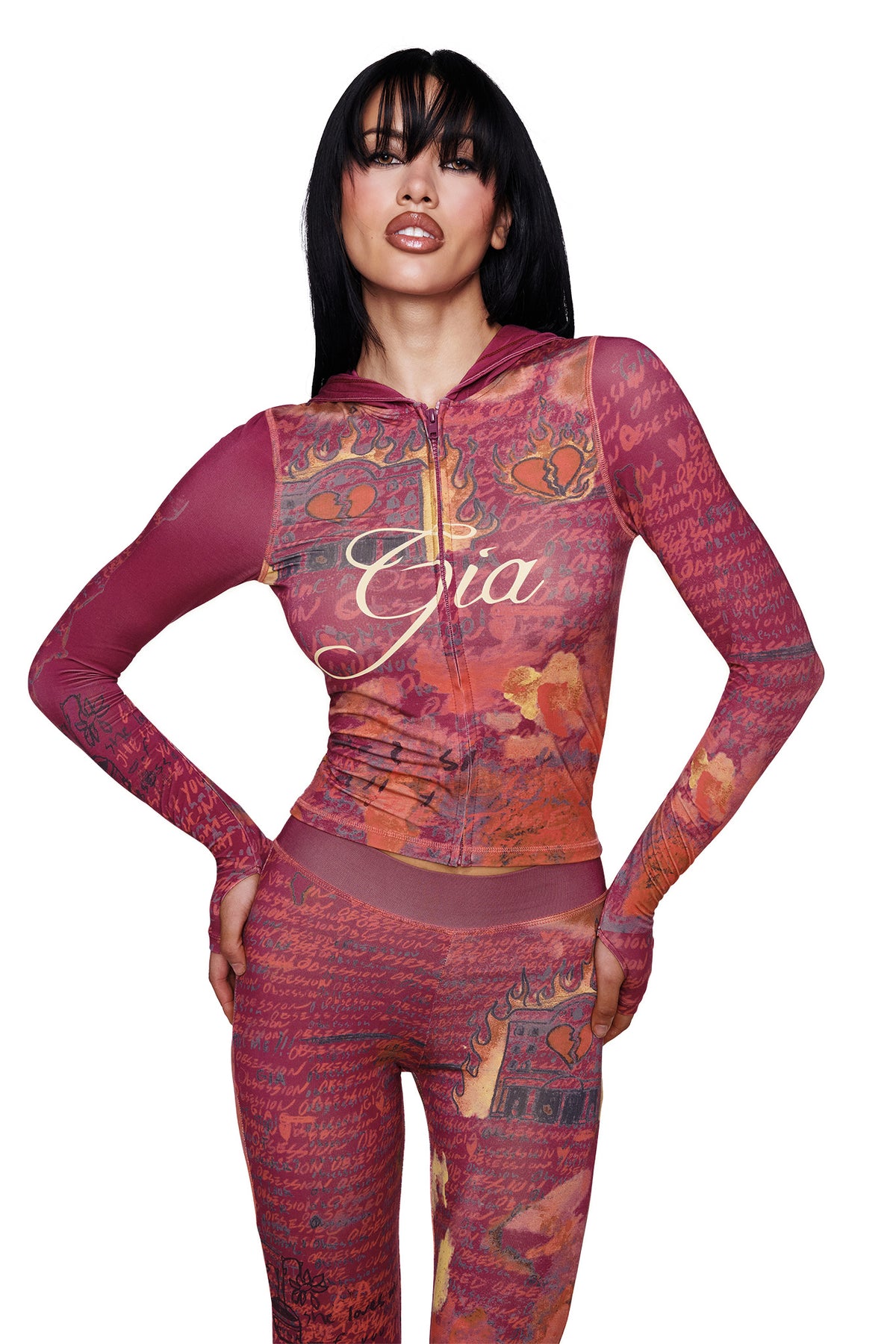 A woman with long black hair poses against a white background, wearing the I.AM.GIA T-REX BLARE HOODIE in red—a fitted jumpsuit featuring maroon and orange patterns, handwritten text, and the I.AM.GIA logo along with "Gia" on the chest. She stands with one hand on her hip.