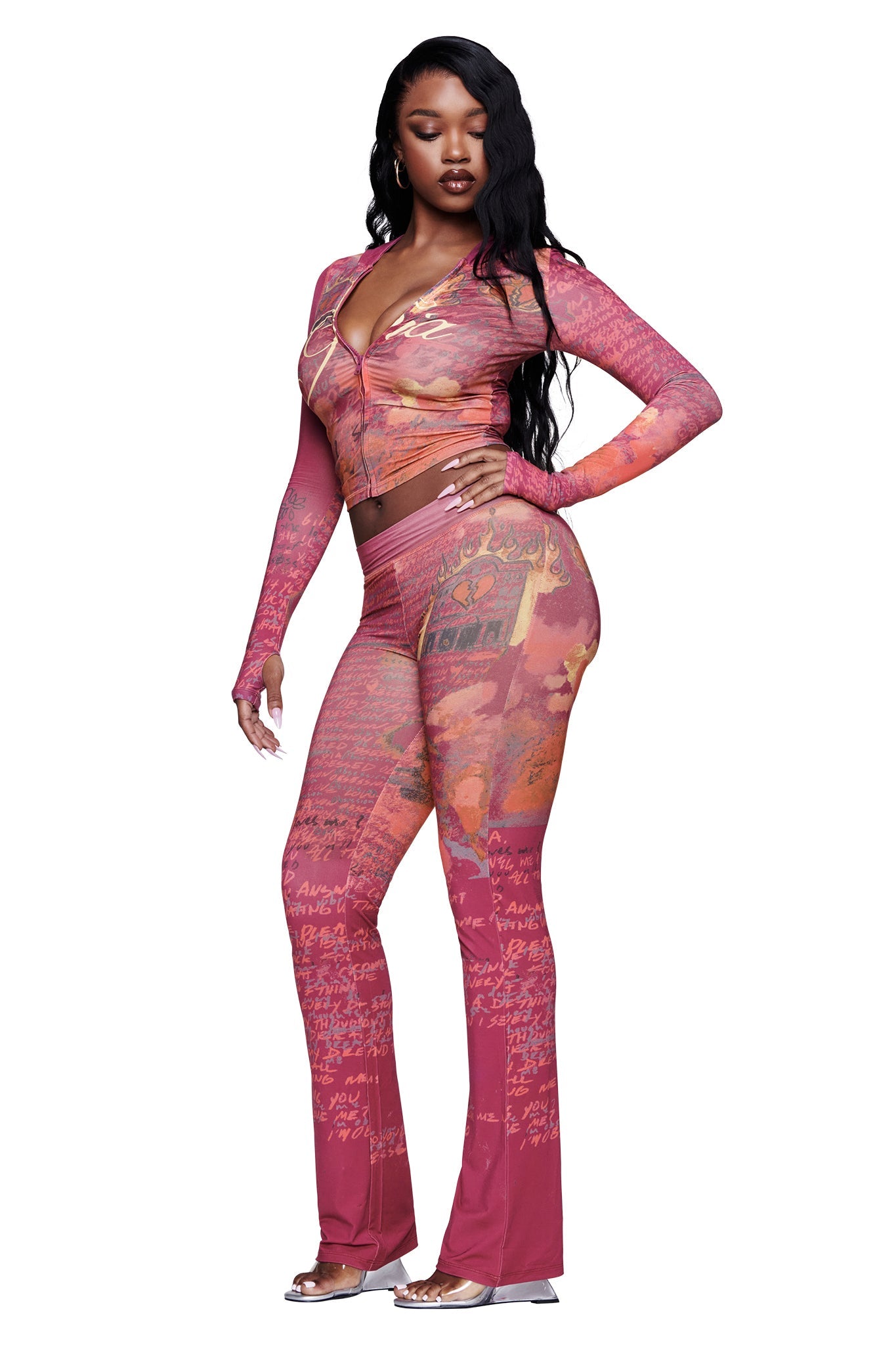 A woman stands in profile against a white background, wearing the I.AM.GIA T-REX BLARE TRACKPANT in red—a fitted, patterned two-piece outfit featuring women's lounge pants with flared legs and long sleeves. She pairs her look with clear high-heeled sandals and wears her long, wavy black hair down.