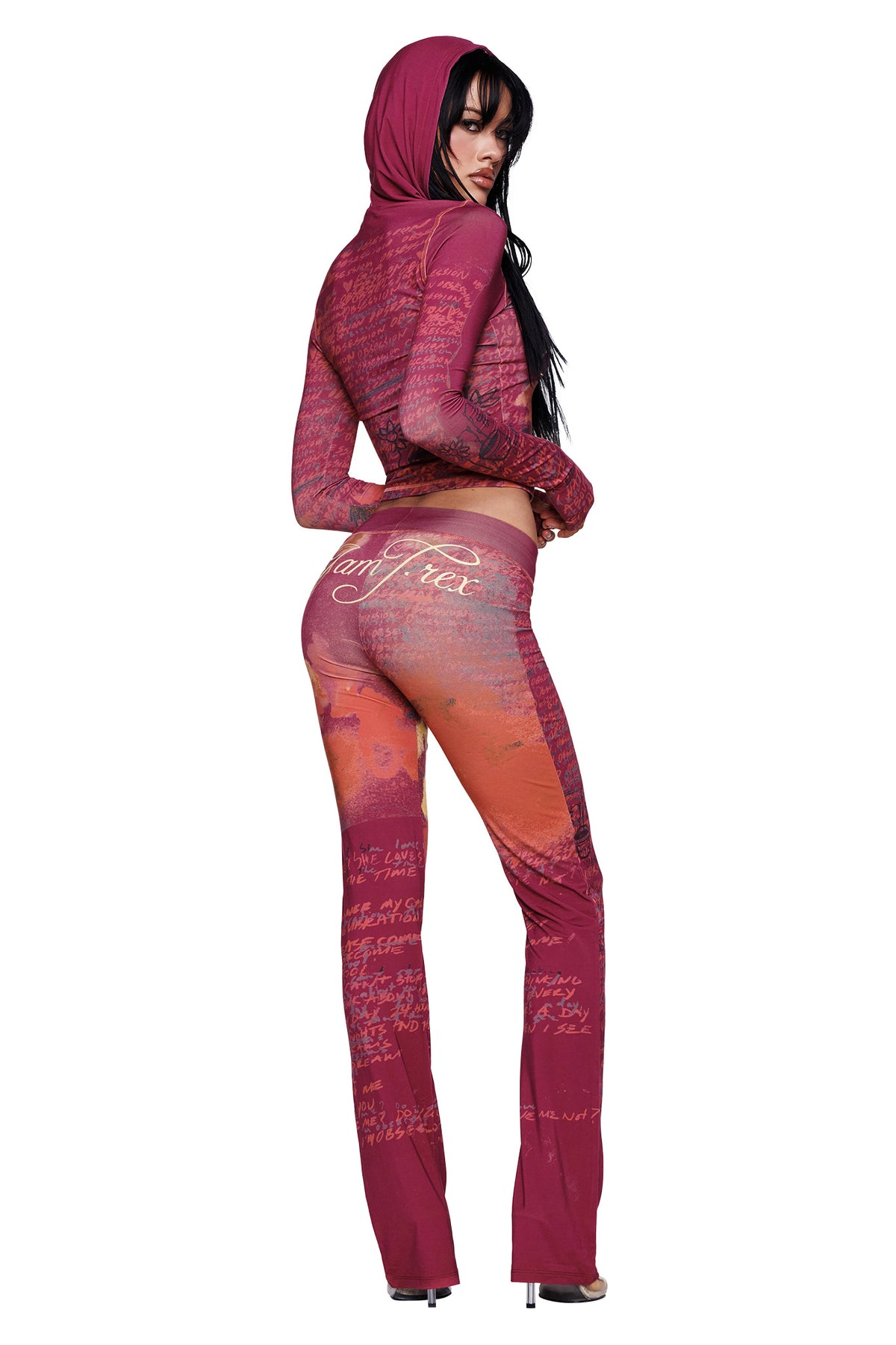 A woman poses with her back turned and glances over her shoulder, wearing high heels and a fitted maroon-and-orange hooded outfit adorned with script text. Her edgy look is highlighted by the I.AM.GIA T-REX BLARE TRACKPANT in bold red.