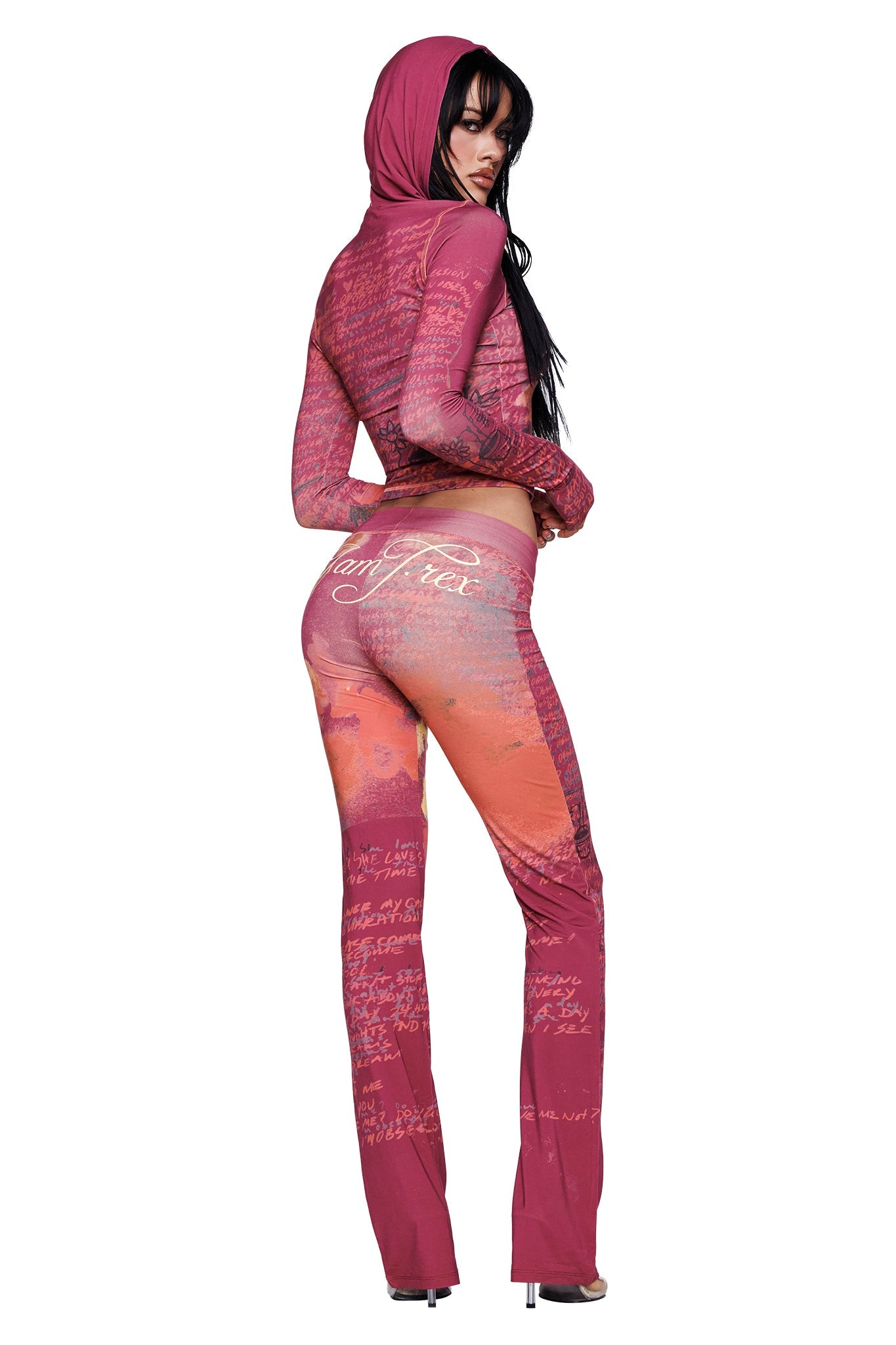 A woman wearing the I.AM.GIA T-REX Blare Trackpant in red, styled with a matching hooded jacket, stands in high heels and looks over her shoulder.