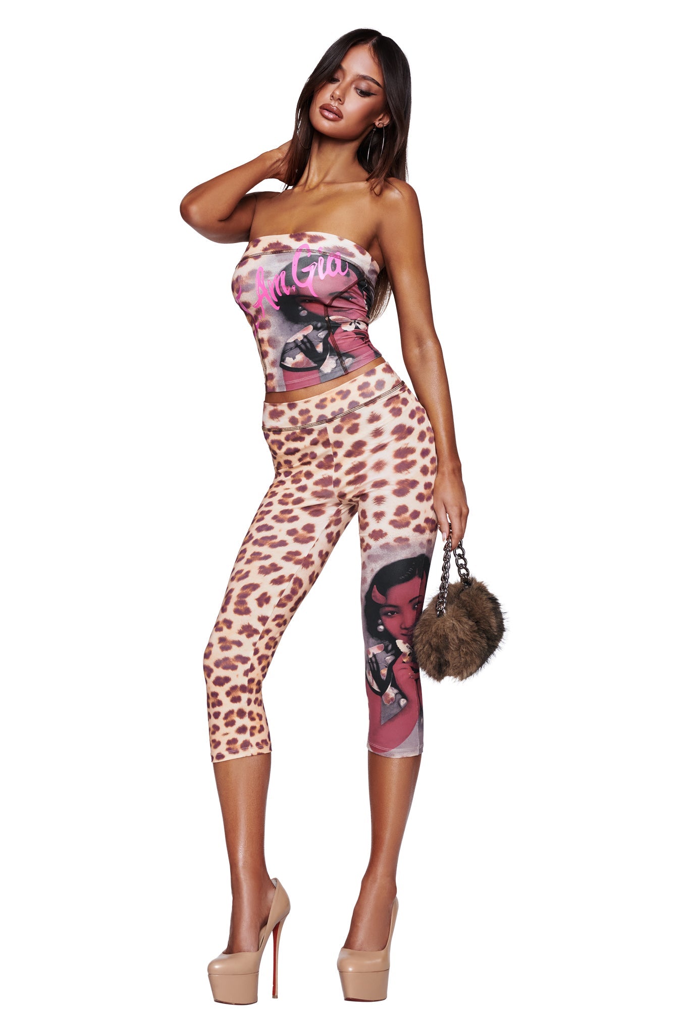 A woman poses in the I.AM.GIA T-REX BLARE BOOB TUBE - WILD CARD and matching leggings with graphic art, holding a fluffy purse. She accessorizes with nude high heels and hoop earrings, standing against a white background. This bold ensemble is perfect for festival style.