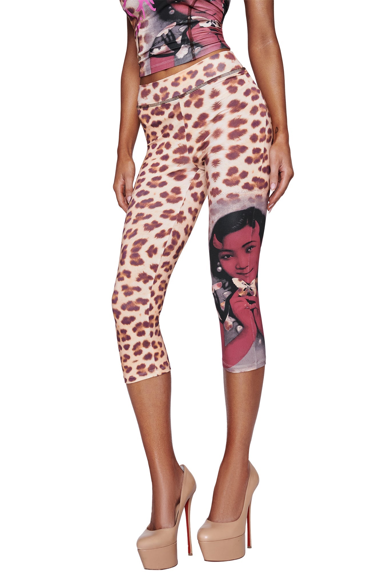 A woman wears the I.AM.GIA T-REX BLARE CAPRI - WILD CARD, featuring cheetah print and a graphic of a woman’s face on one leg. She pairs them with a patterned top and nude platform heels, photographed against a white background.