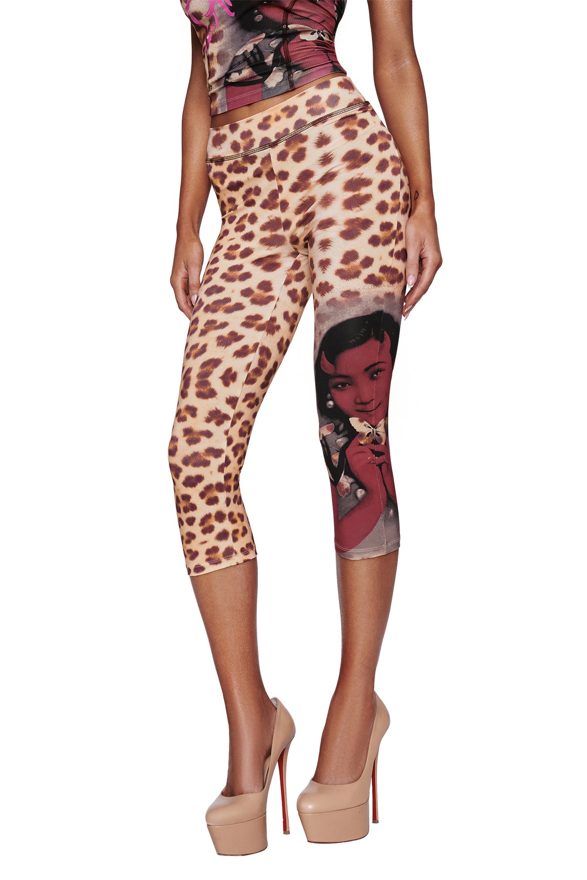 A woman poses confidently against a white background wearing the I.AM.GIA T-REX BLARE CAPRI - WILD CARD, featuring a cheetah print and a graphic of a woman's face on one leg, paired with nude high heels.