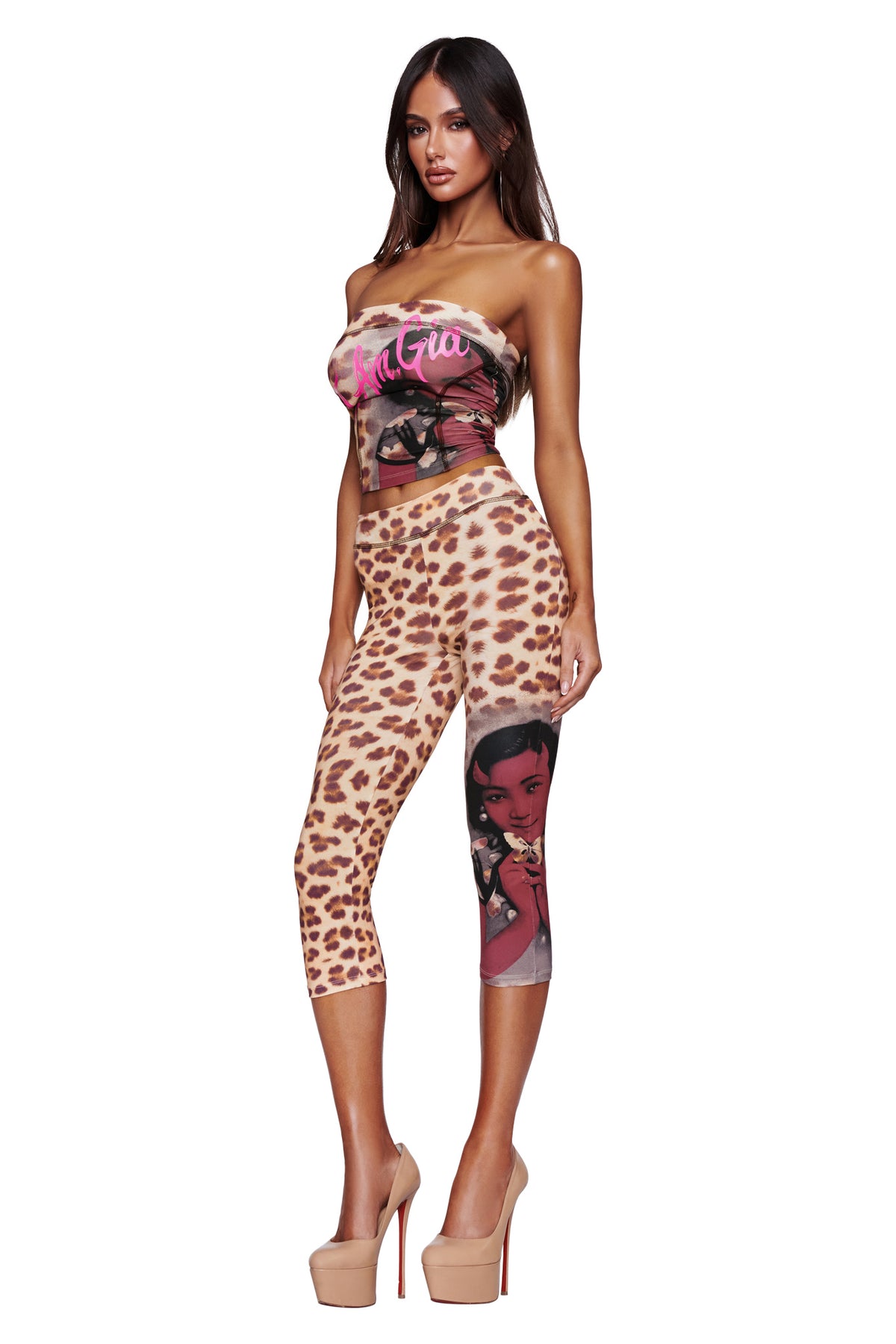 A woman stands against a white background wearing the I.AM.GIA T-REX BLARE BOOB TUBE - WILD CARD, styled with high heels and leopard print leggings featuring a cartoon woman illustration on one leg.