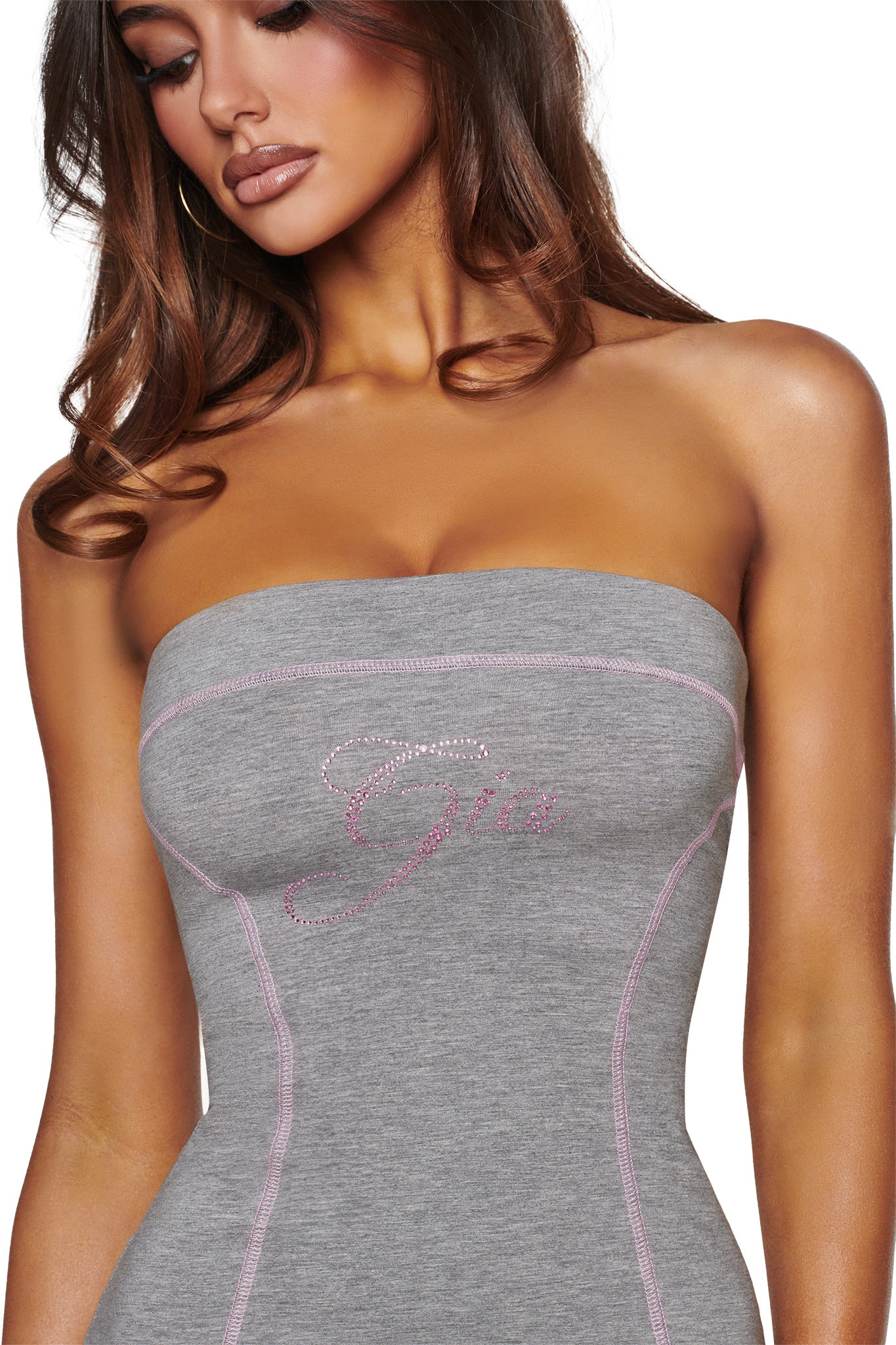 A woman wears the I.AM.GIA BLARE TUBE MINI DRESS in heather grey, featuring pink stitching and “Gia” spelled out in pink rhinestones across the chest. Her loose, wavy brown hair frames her partially visible face.