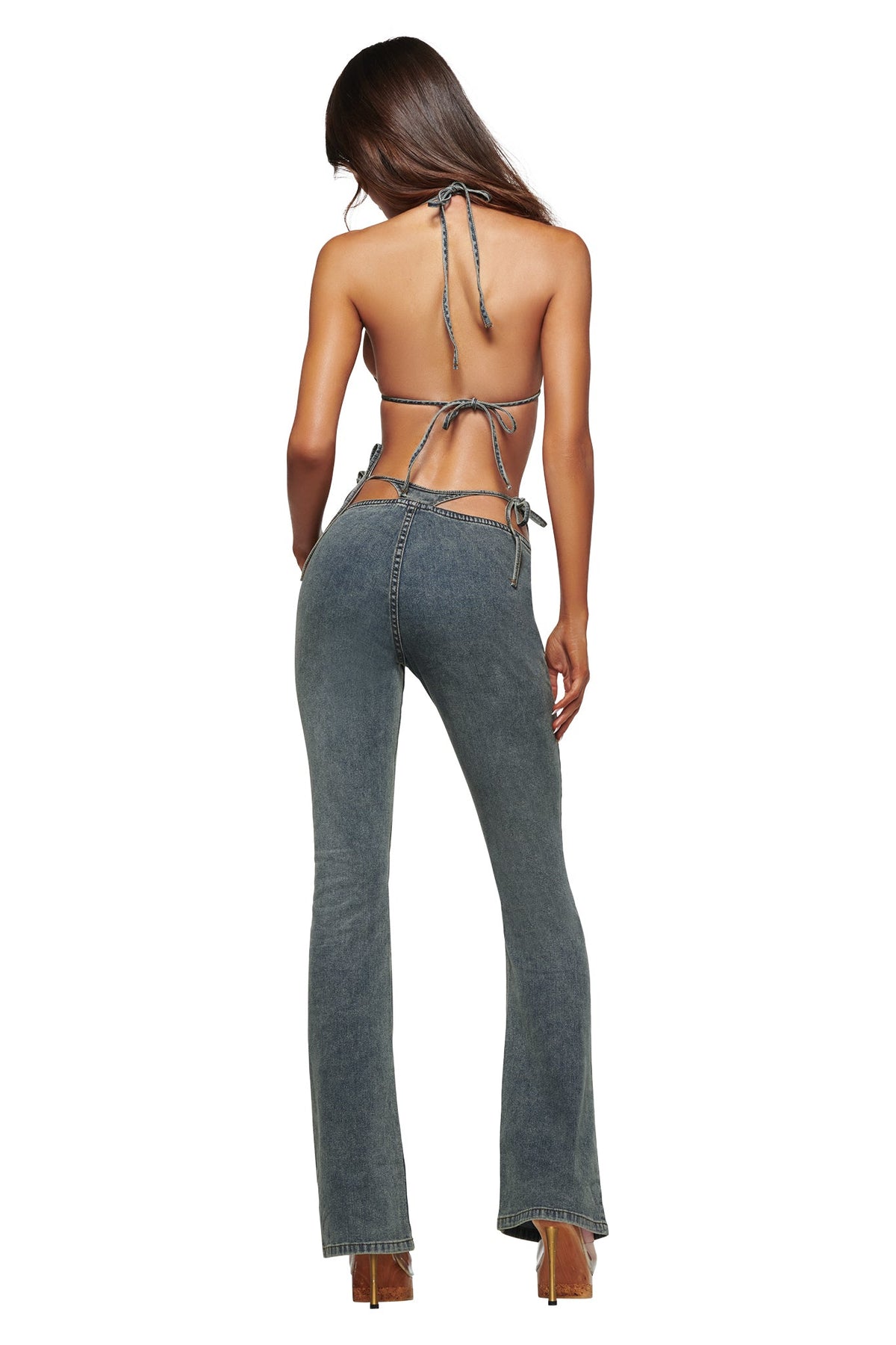 A woman stands with her back to the camera against a white background, wearing the I.AM.GIA JET G-STRING DENIM PANT in faded indigo—high-waisted, flared blue jeans with chain details inspired by iconic Y2K style—paired with a backless, strappy halter top.