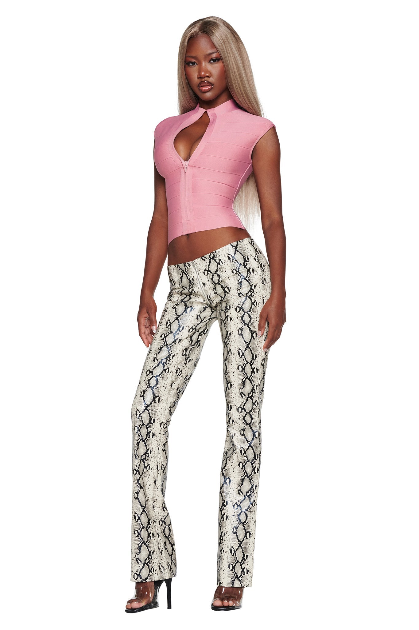 A woman stands against a white background wearing a pink sleeveless top with a front cutout, styled with the I.AM.GIA HENDRIX 2.0 PANT in Cream Snake—a pair of high-waisted, straight-leg faux python leather pants—paired with black high-heeled sandals for a chic night-out look.