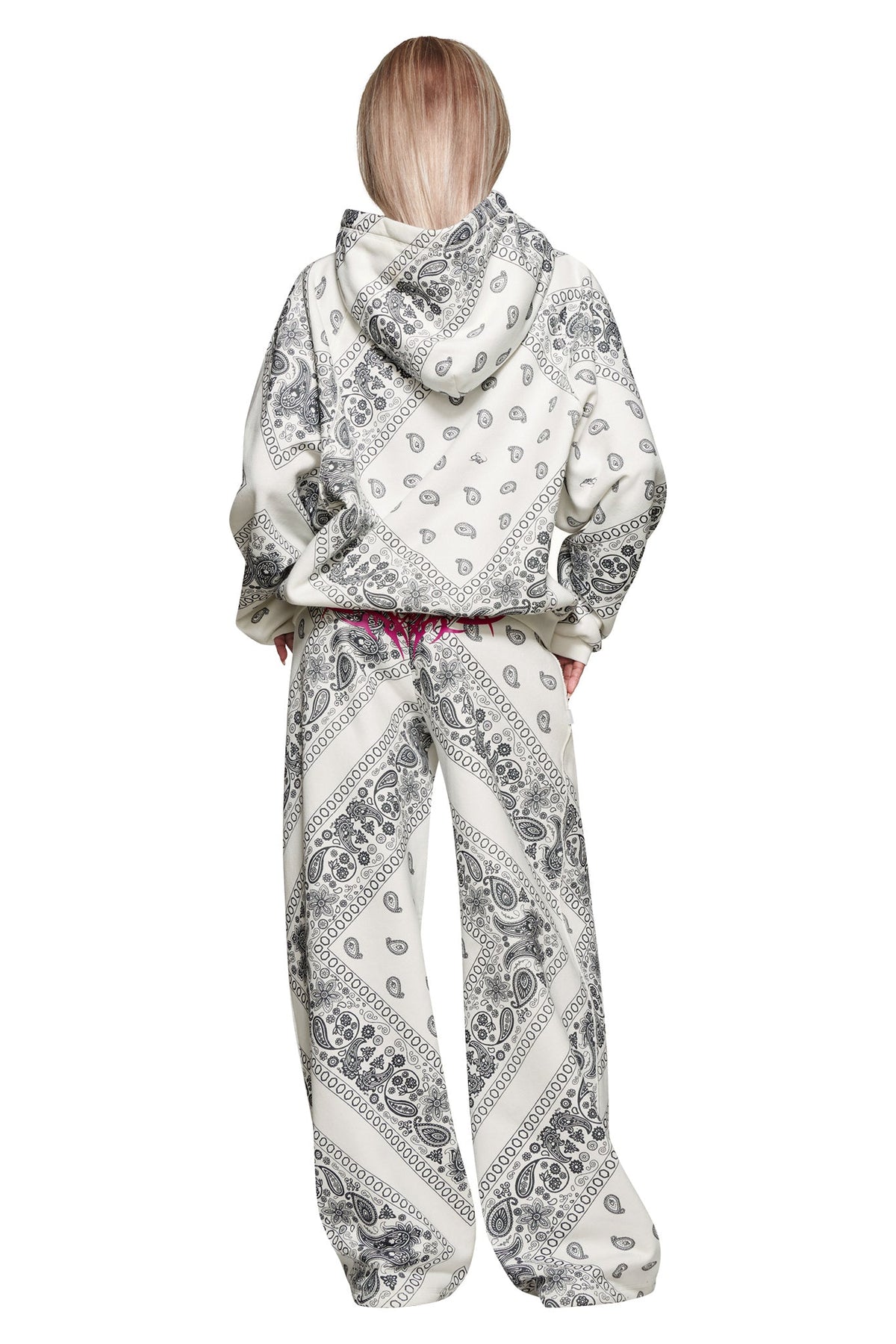 A person stands with their back to the camera, hands in pockets, wearing I.AM.GIA’s T-REX TRACKPANT - PAISLEY in white with a gray paisley bandana pattern. Long straight hair worn loose. Part of the T-REX collaboration.