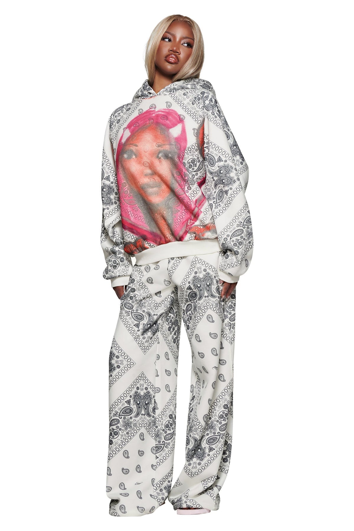A woman wears I.AM.GIA’s T-REX TRACKPANT - PAISLEY and a matching white bandana-print hoodie with a bold graphic of a woman with bright pink hair and nails, standing against a white background.