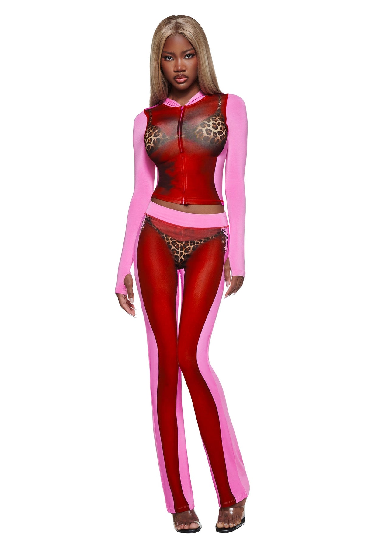 A woman models the I.AM.GIA T-REX SPRAY BODY BLARE HOODIE - PINK, a sheer red and pink piece with leopard print accents, long sleeves, and an airbrushed bikini body print. She pairs it with high-heeled sandals.