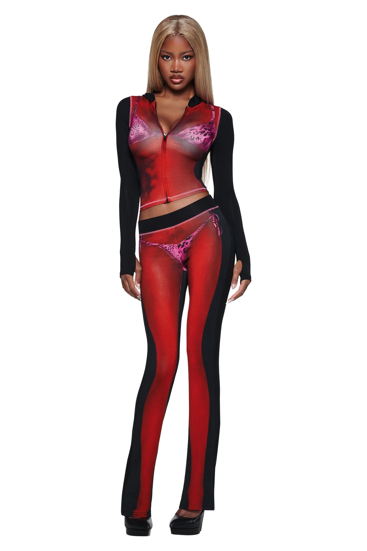 A woman with long blonde hair wears a fitted, sheer-effect outfit and the T-REX SPRAY BODY BLARE TRACKPANT - BLACK by I.AM.GIA, perfect for standout airport style. She stands facing forward.