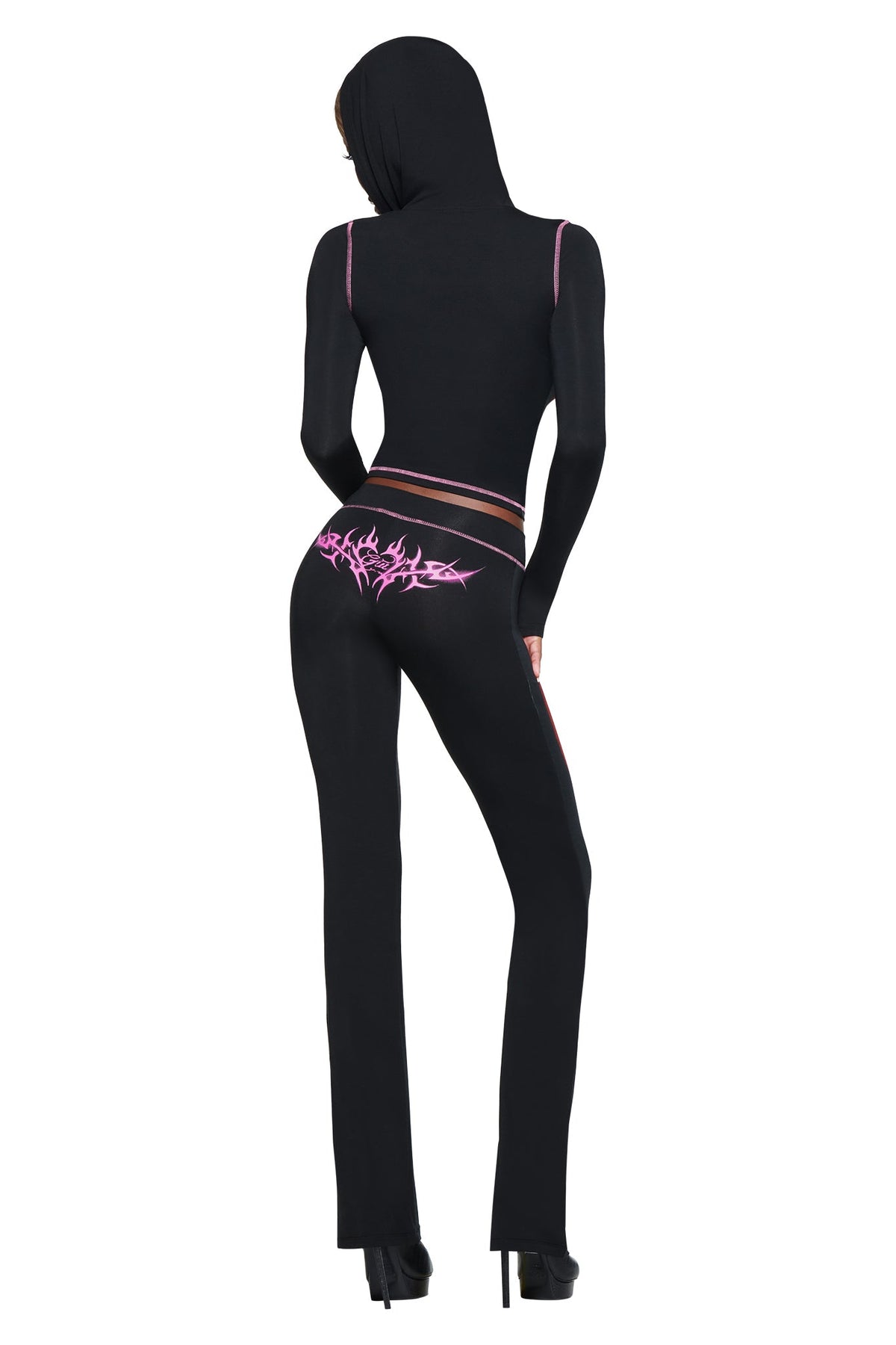 Person wearing a fitted black hooded outfit with pink accents and T-REX SPRAY BODY BLARE TRACKPANT - BLACK by I.AM.GIA, back to camera. Pink graphic detail on lower back. High heels complete the airport styling look.