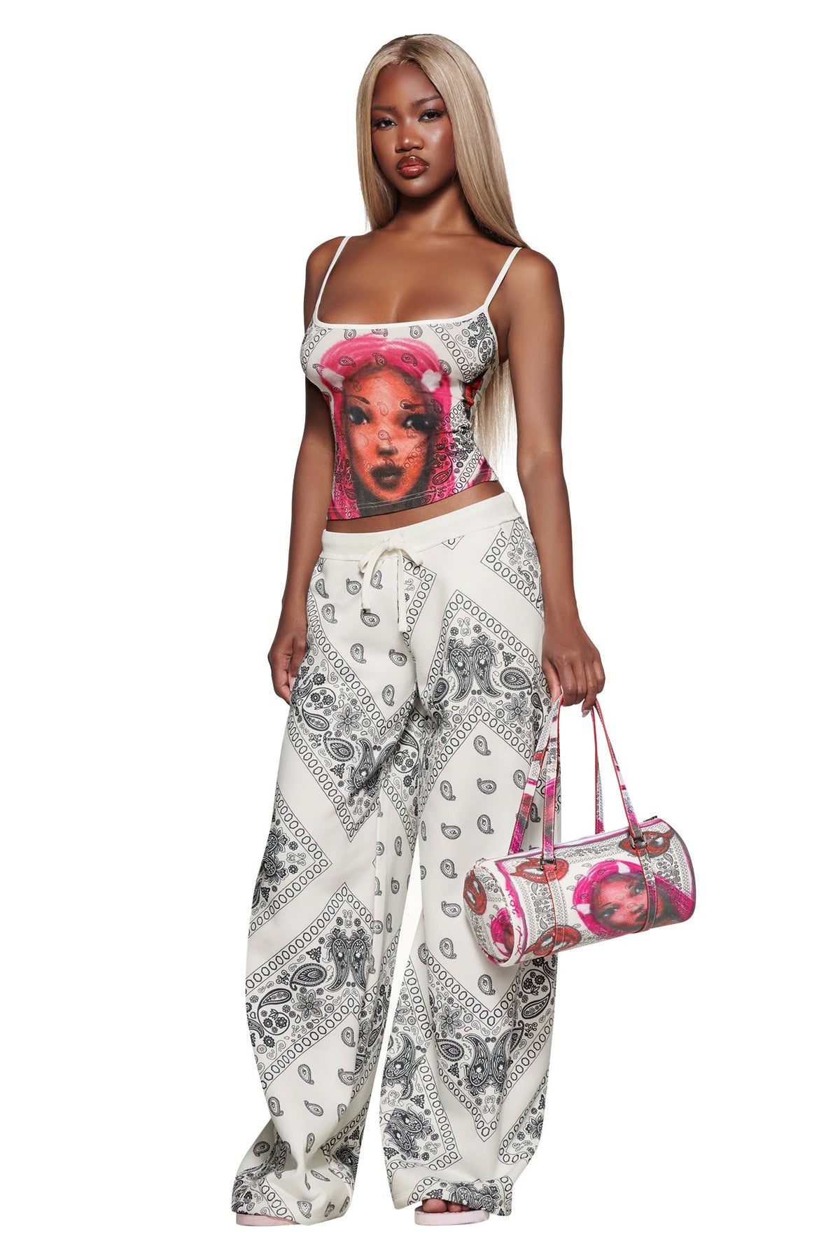 A woman with long blonde hair wears the I.AM.GIA T-REX TANK - PAISLEY FACE, white bandana-print wide-leg pants, and holds a pink patterned handbag while standing against a white background.