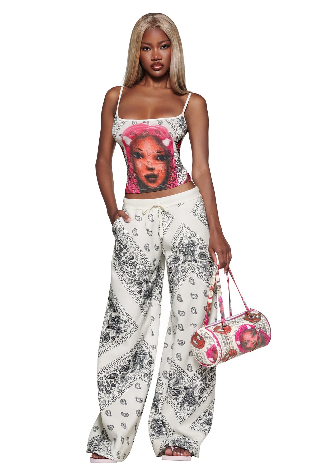 A woman with long blonde hair wears the I.AM.GIA T-REX TANK - PAISLEY FACE, a crop-length white tank with a bold red woman’s face graphic, paired with wide-leg bandana print pants and a matching bag.