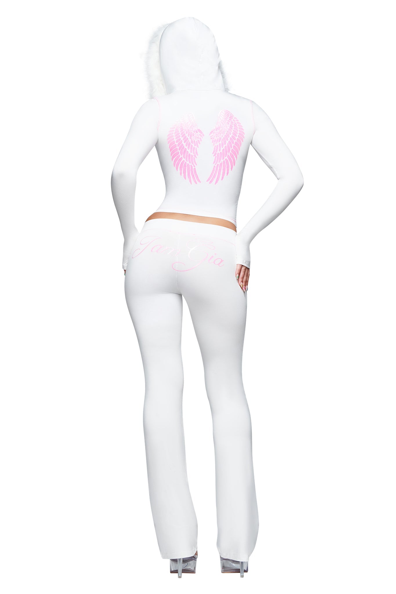 A person stands with one hand on their hip and high heels, photographed from behind, wearing the I.AM.GIA BLARE TRACKPANT - WHITE : ANGEL. This bamboo trackpant features a white hooded outfit with pink angel wings graphic on the back, text detailing on the pants, a low-rise waist, and sleek flared legs for a modern silhouette.