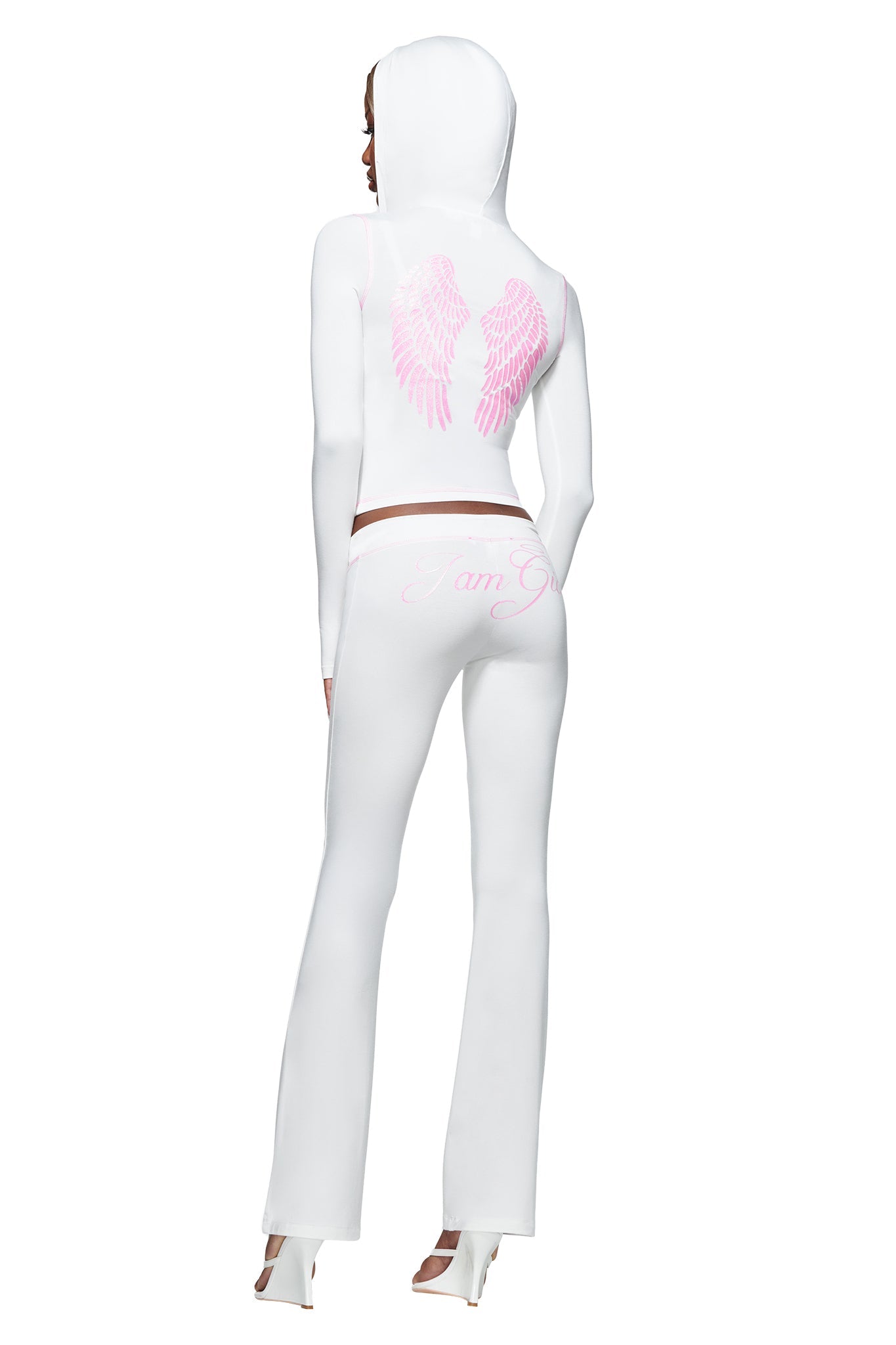 A woman stands facing away, wearing the I.AM.GIA BLARE FUR TRIM HOODIE in white, which features pink wing graphics on the back. She pairs it with matching pants with pink script and high heels, adding a touch of celestial charm to her stylish look.