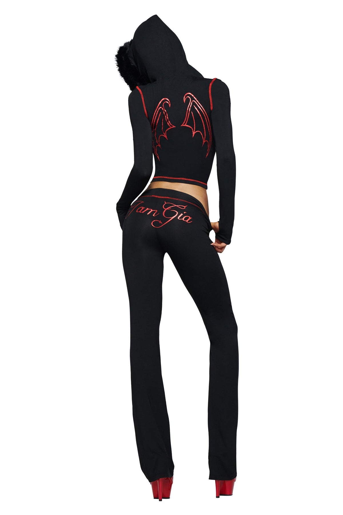 A person, seen from behind with hands on hips, sports the I.AM.GIA BLARE TRACKPANT - JET BLACK : DEVIL featuring a bold black devil GIA logo and the “I.AM.GIA” branding, paired with a black hooded outfit adorned with striking red bat wings and finished off with red high heels.