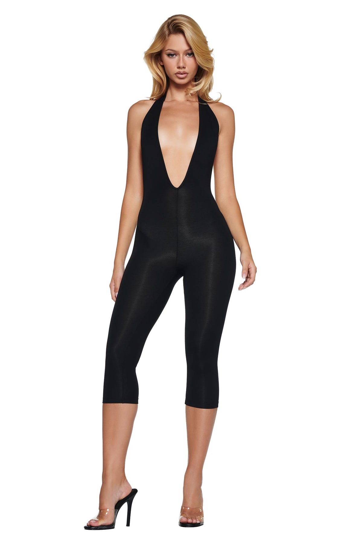 A model wears the I.AM.GIA BLARE HALTER CAPRI JUMPSUIT - BLACK, a black halter-neck capri jumpsuit with a deep neckline and clear high-heeled sandals.
