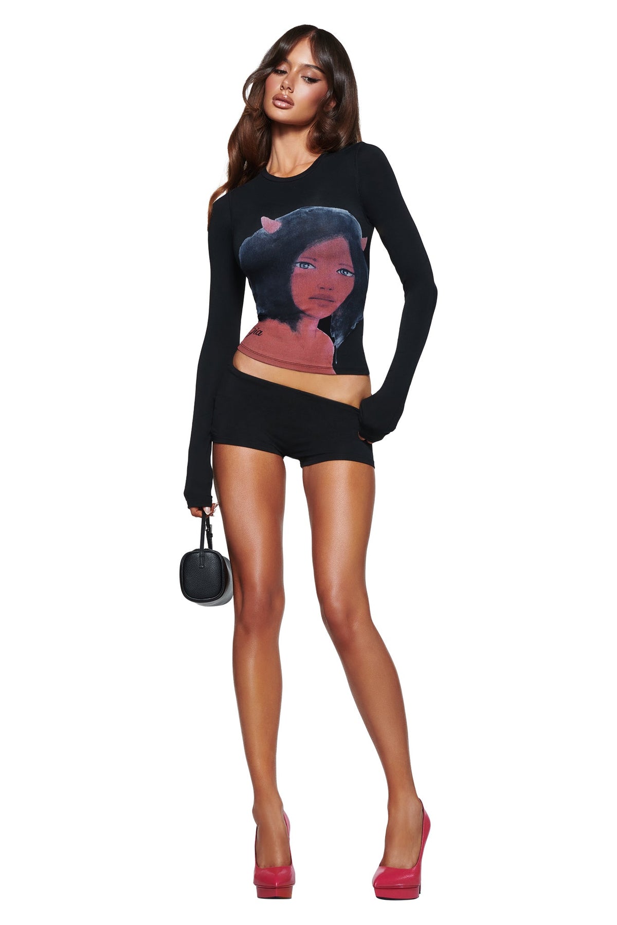 A woman wears the I.AM.GIA T-REX BLARE 2.0 LONG SLEEVE TEE - BLACK with black shorts, pink high heels, and a small black purse while posing against a white background.