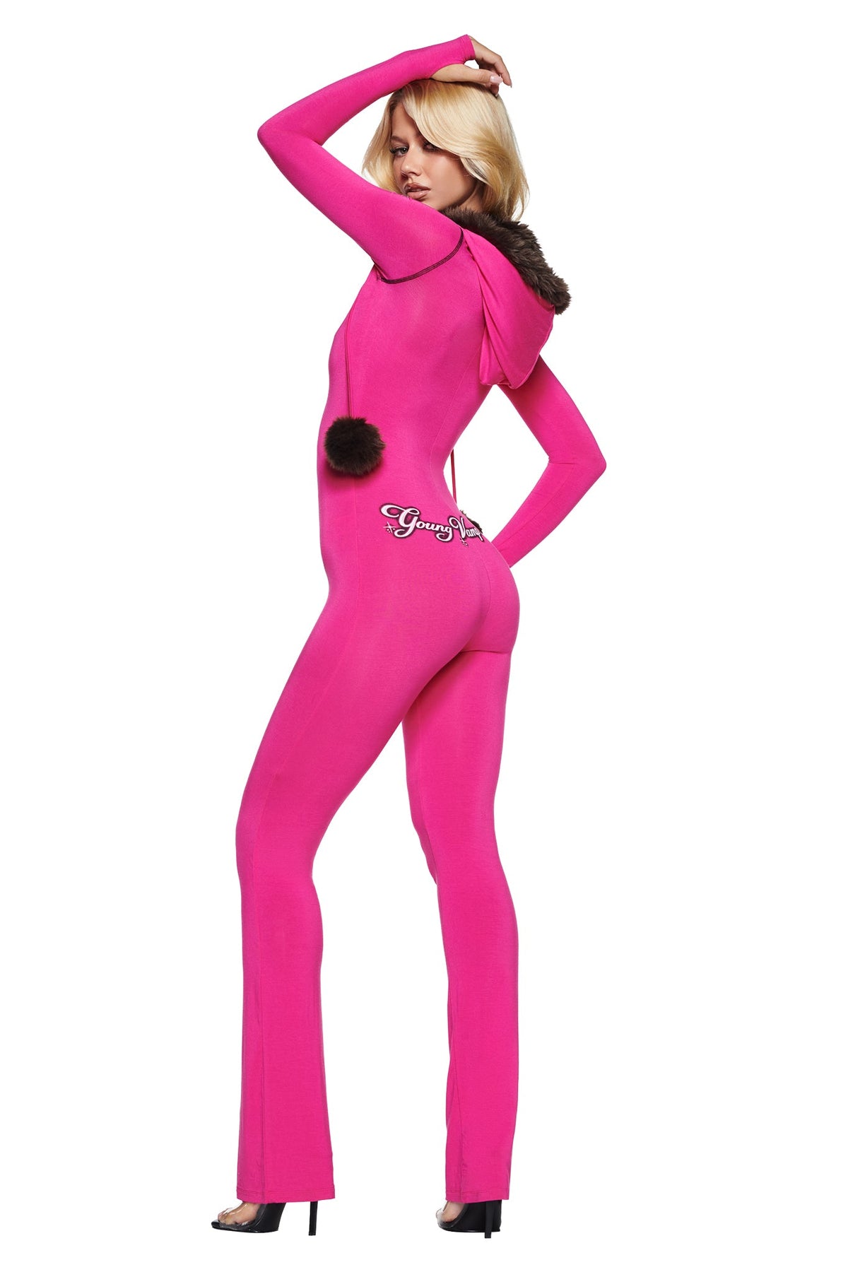A woman wears a pink YVL BLARE FUR LINED JUMPSUIT, a long-sleeved, faux fur hooded jumpsuit with a dark pom-pom detail on the back.