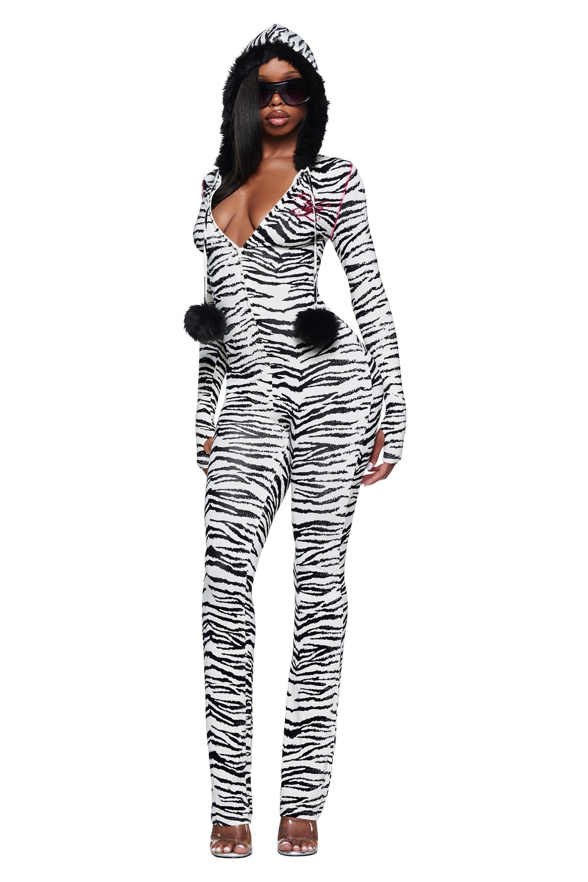 A person wears a long-sleeved I.AM.GIA YVL BLARE FUR LINED JUMPSUIT - ZEBRA, featuring a faux fur hood and black pom-poms hanging from the front zipper.