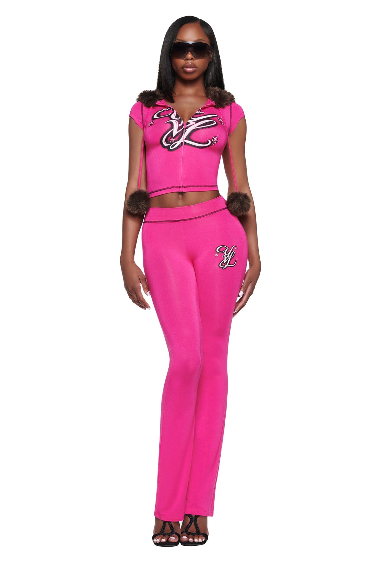 A person wears a hot pink zip-up cropped top with fur-trimmed hood strings and matching high-waisted YVL BLARE TRACKPANT - PINK, both featuring black and white graphic logos and a flared leg.