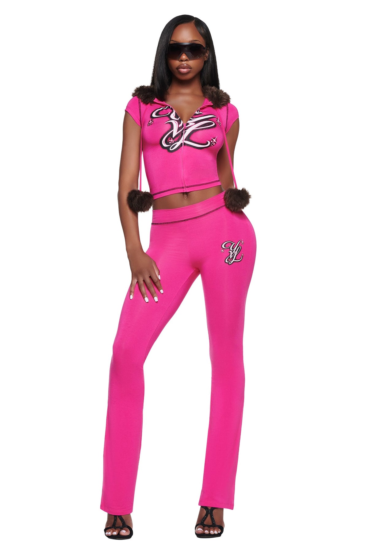 A model wears a bright pink matching set featuring a zippered, short-sleeve hooded crop top with fur trim and pom-poms, paired with I.AM.GIA YVL BLARE TRACKPANT - PINK flared trousers with embroidered logos.