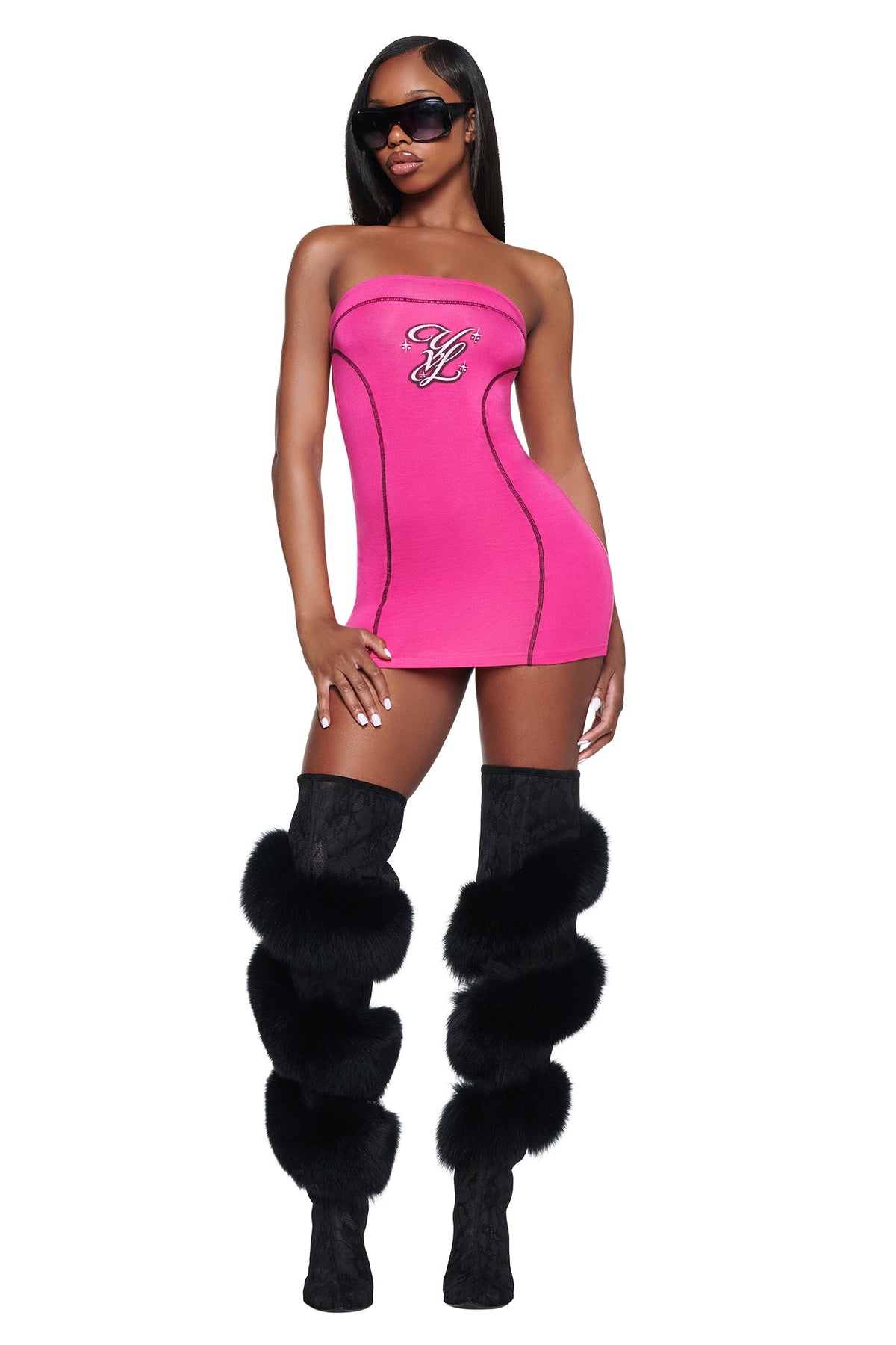 A person wears a hot pink I.AM.GIA YVL BLARE TUBE MINI DRESS - PINK featuring black stitching and a chest logo, paired with black thigh-high boots adorned with large fur pom-poms.